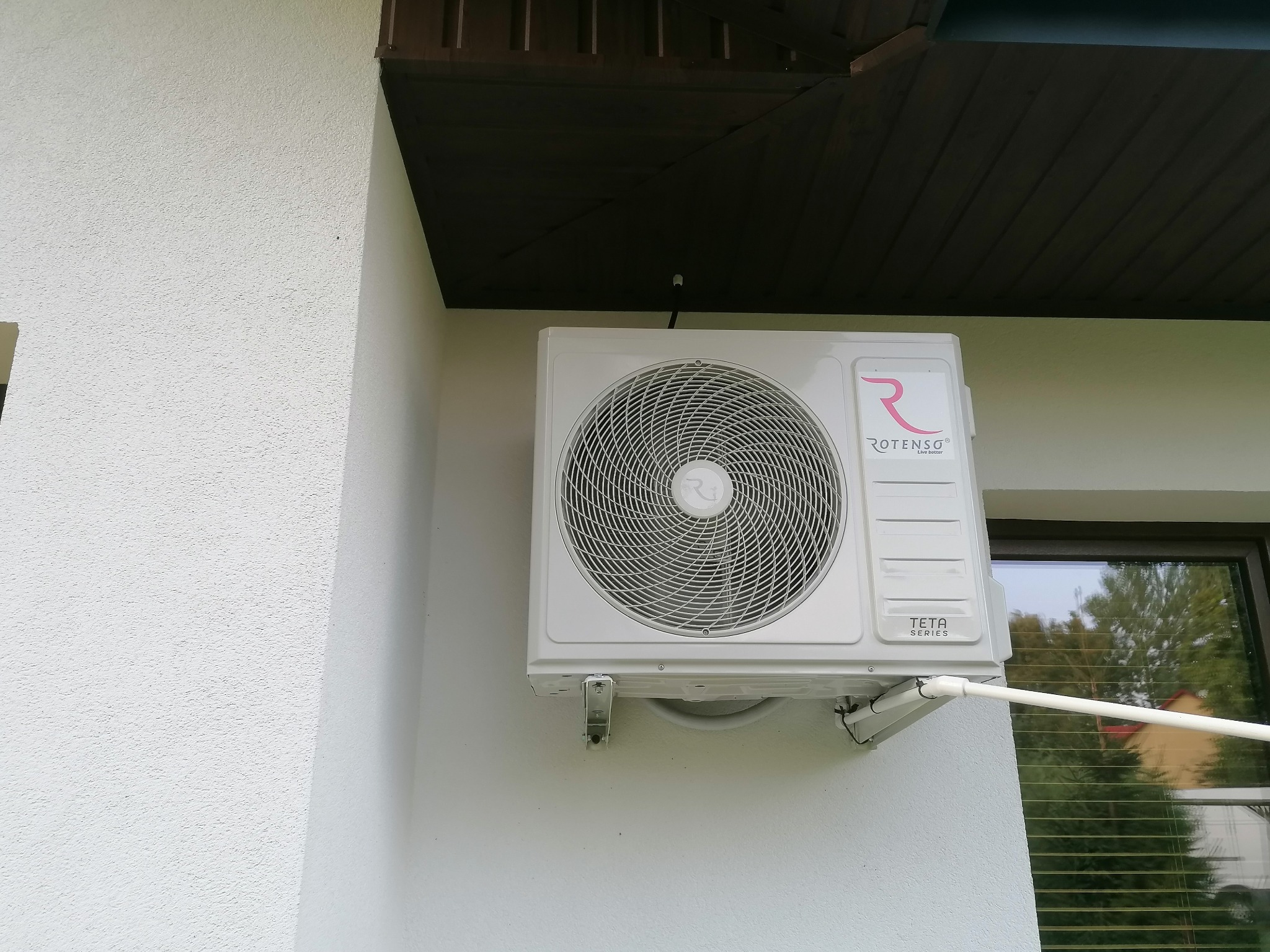 Biała jednostka zewnętrzna klimatyzacji Rotenso Teta Series zamontowana na elewacji budynku, widoczny fragment okna i brązowe podbitki dachu.