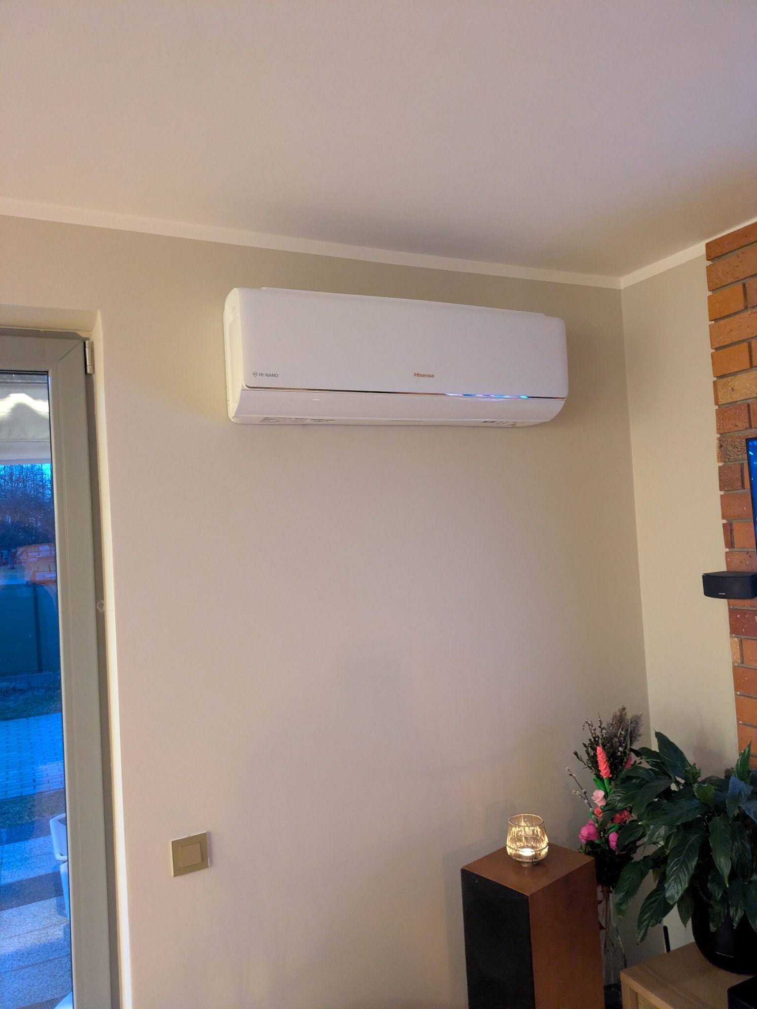 Biały klimatyzator Hisense zamontowany na ścianie w jasnym wnętrzu. Widoczny fragment ceglanej ściany i dekoracje roślinne. Subtelne oświetlenie LED pod urządzeniem.