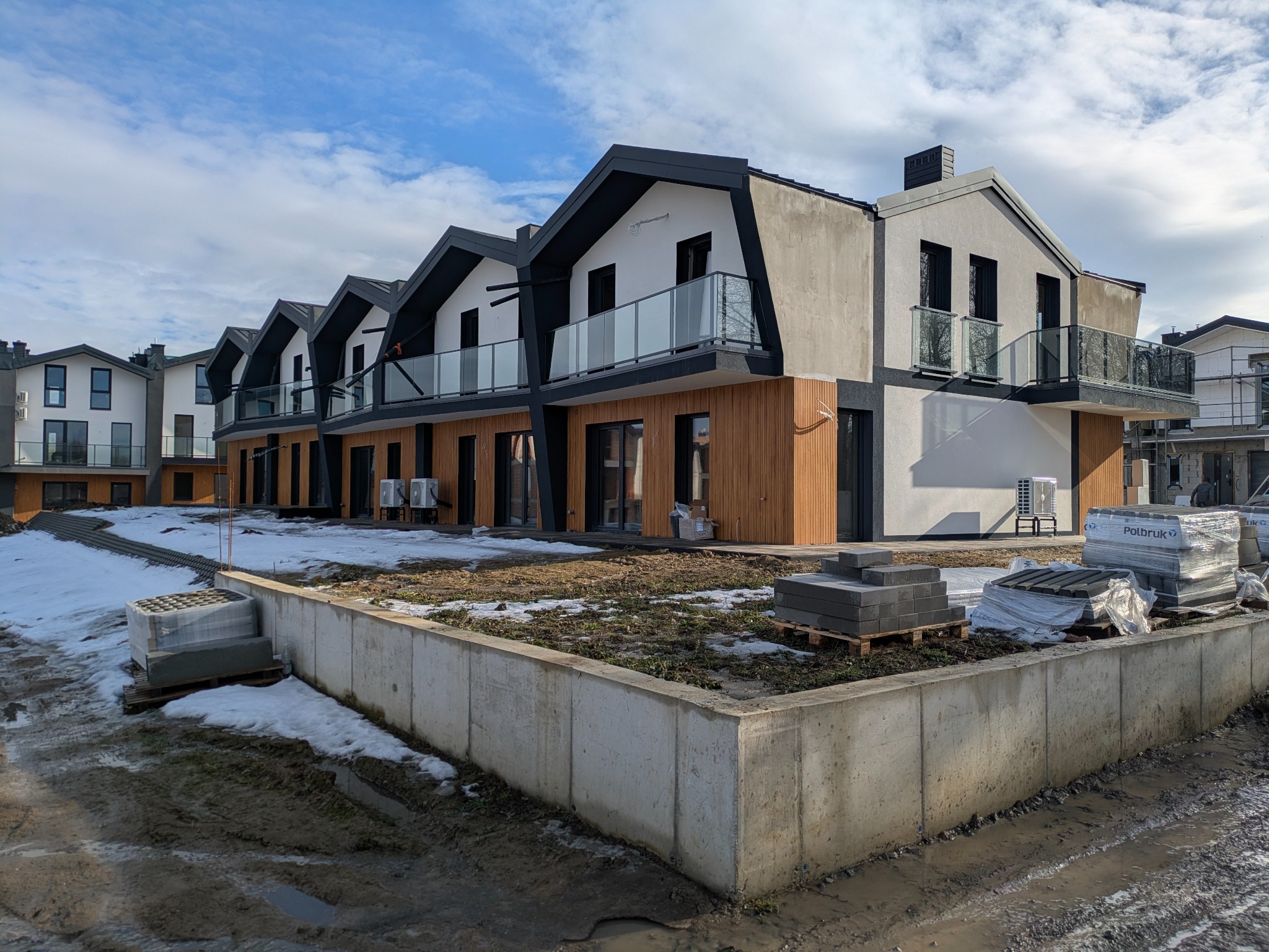 Nowoczesne domy szeregowe z balkonami i klimatyzacją zewnętrzną, w trakcie wykończenia. Widoczne materiały budowlane i fragmenty topniejącego śniegu.