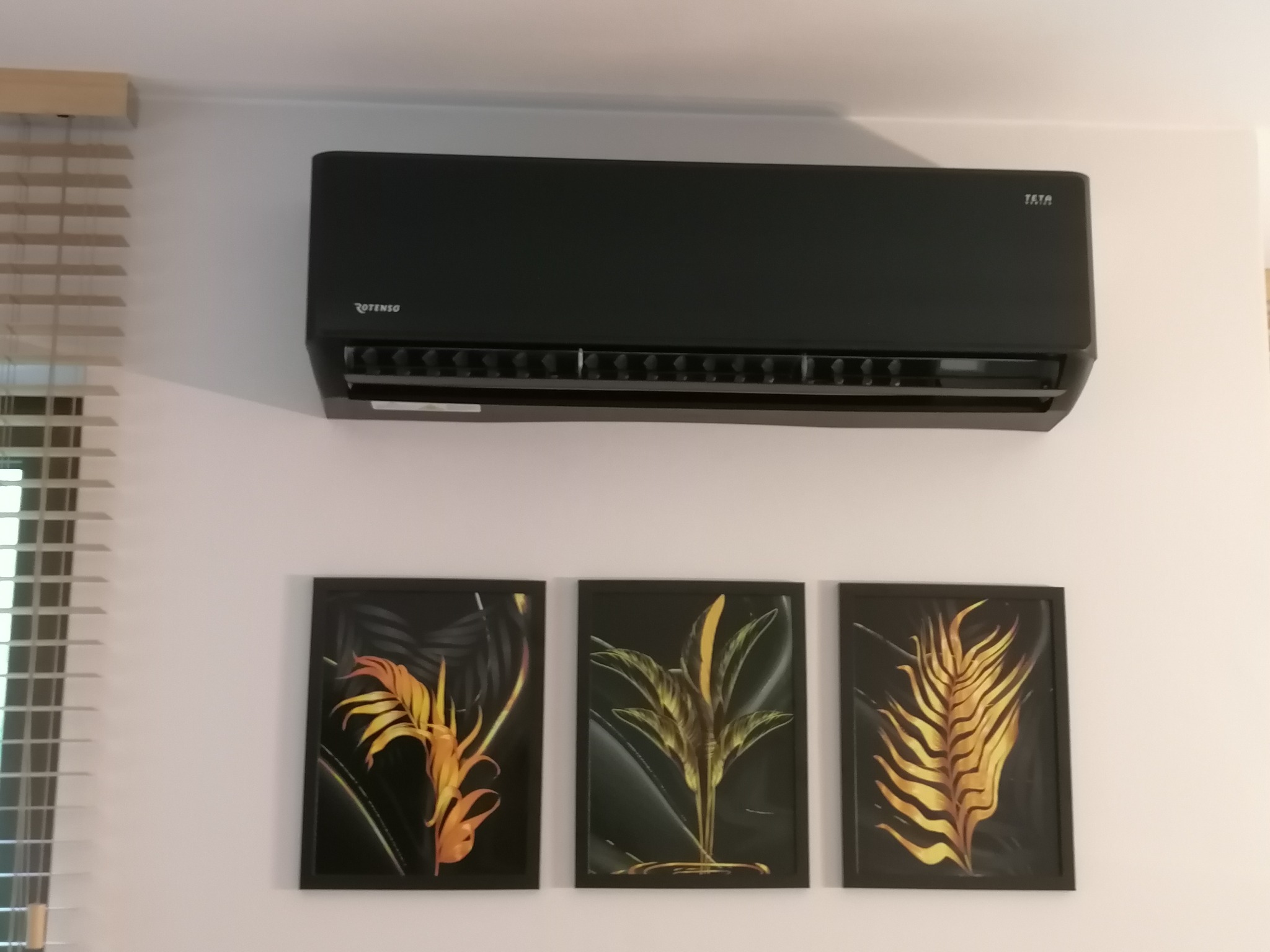 Czarny klimatyzator Rotenso, model TATA, zamontowany na białej ścianie nad trzema obrazami z motywami roślinnymi. Widoczne żaluzje okienne po lewej stronie.