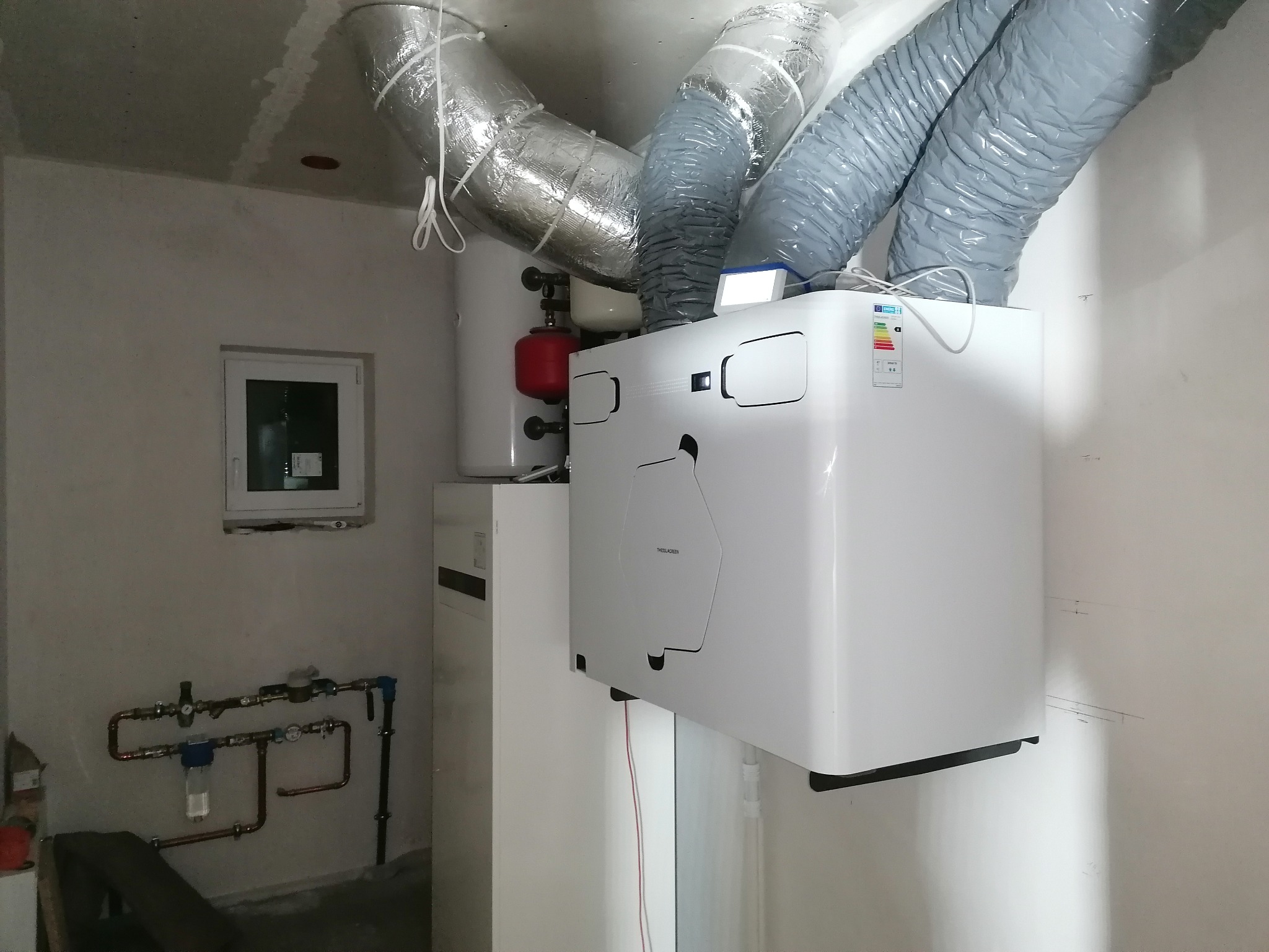 Biała centrala wentylacyjna z podłączonymi przewodami wentylacyjnymi w surowym pomieszczeniu. Widoczne rury i instalacja wodna. Małe okno w tle.