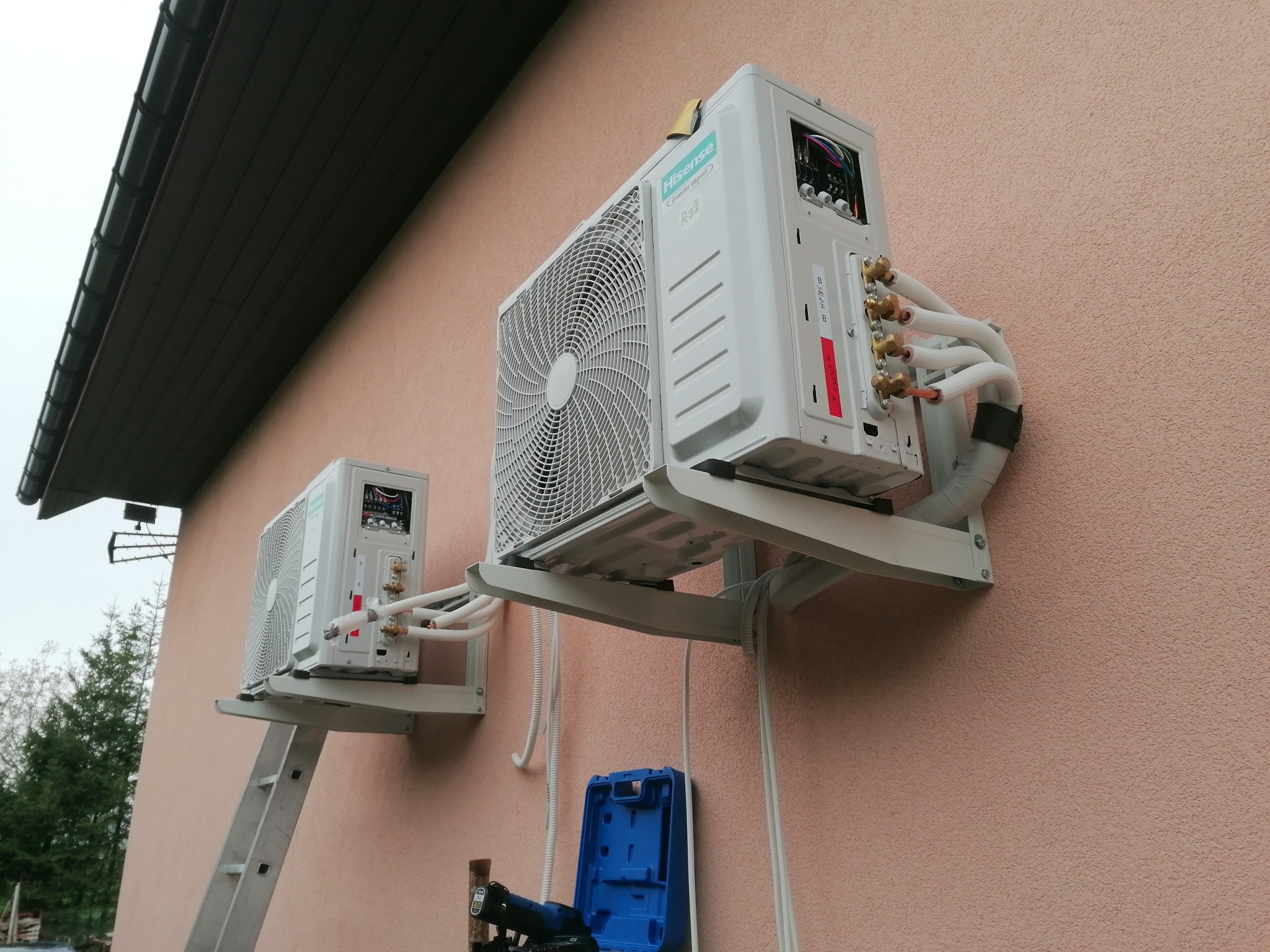 Dwa białe klimatyzatory Hisense zamontowane na elewacji budynku, widoczne rury i narzędzia instalacyjne. Jeden klimatyzator na drabinie.
