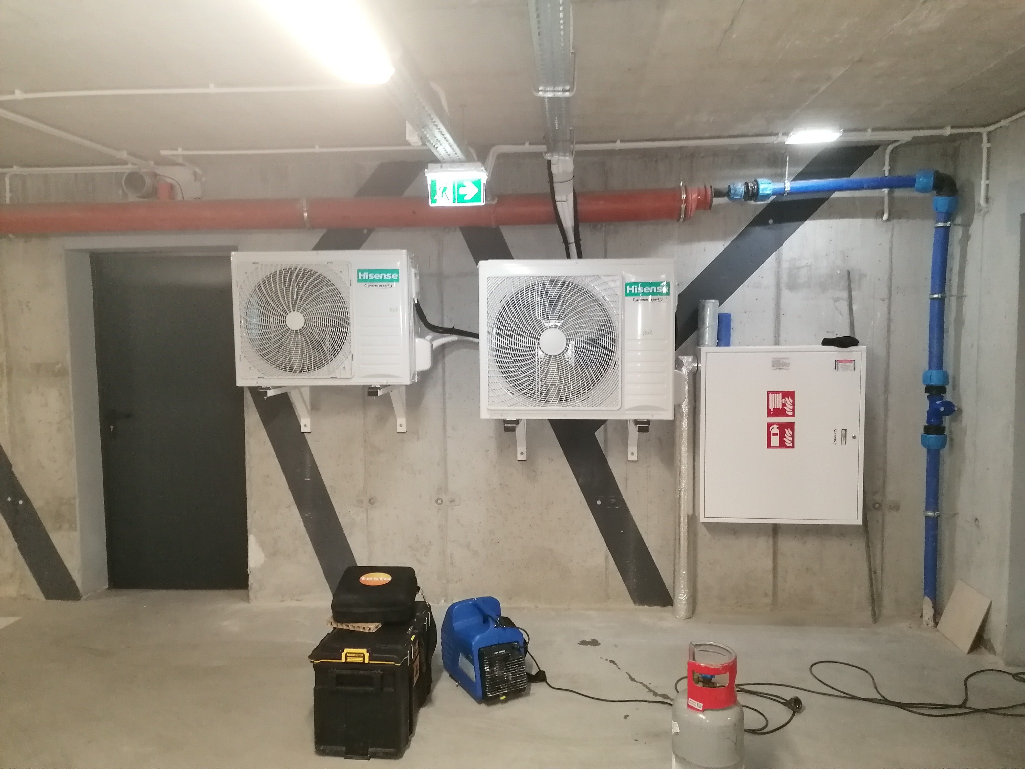 Dwa białe klimatyzatory Hisense na betonowej ścianie w garażu, z widocznymi rurami, skrzynką przeciwpożarową i narzędziami, pod zielonym znakiem ewakuacyjnym.