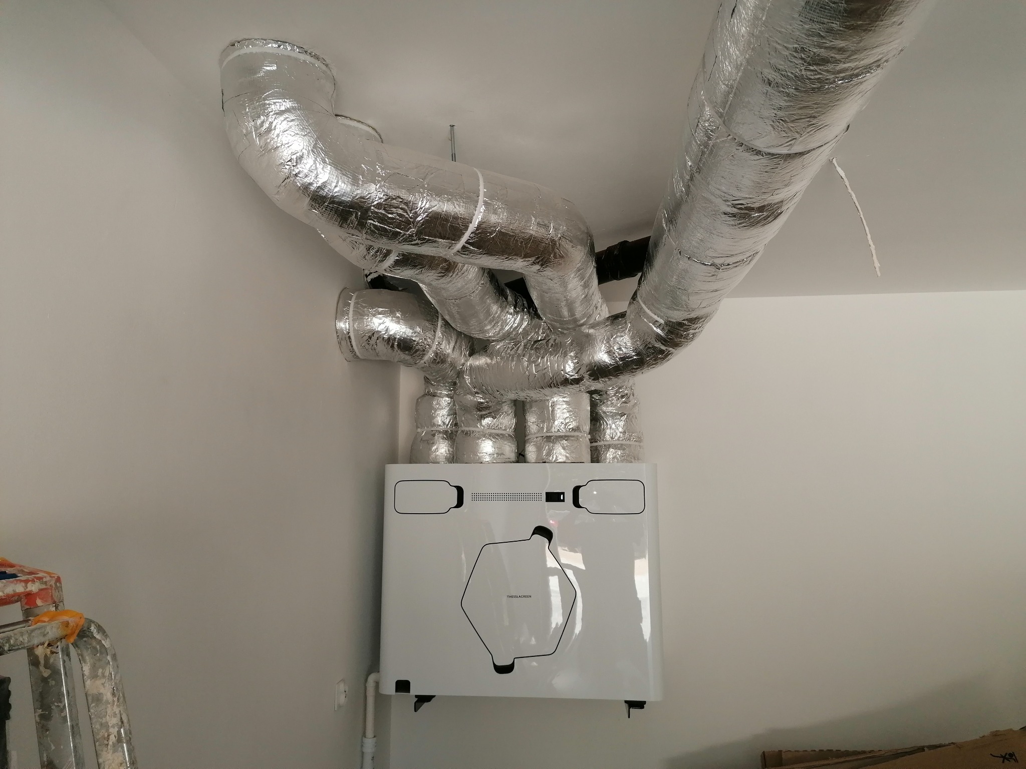 Biała centrala wentylacyjna z podłączonymi, izolowanymi przewodami wentylacyjnymi w srebrnej folii, zamontowana na ścianie w jasnym pomieszczeniu. Widoczny fragment drabiny.