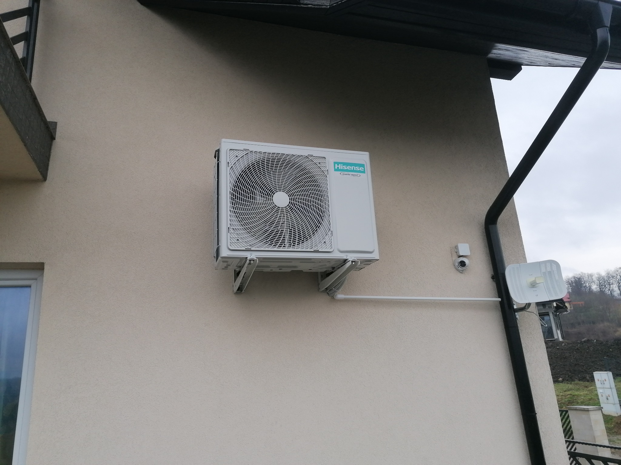 Biała jednostka zewnętrzna klimatyzacji Hisense zamontowana na elewacji budynku z beżowym tynkiem, widoczna rynna i kamera monitoringu. Ujęcie z dołu.