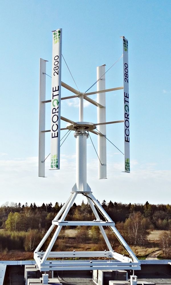 Biała turbina wiatrowa pionowa z napisem 'ECOROTE 2800' na tle błękitnego nieba i drzew. Widok z dołu, ukazujący konstrukcję wsporczą i detale turbiny.