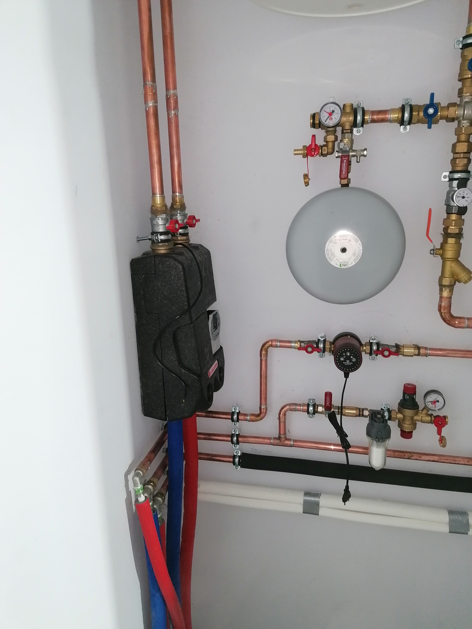 Zamknięta instalacja grzewcza z czarnym, izolowanym wymiennikiem ciepła, miedzianymi rurami, manometrami i zbiornikiem wyrównawczym na białej ścianie.