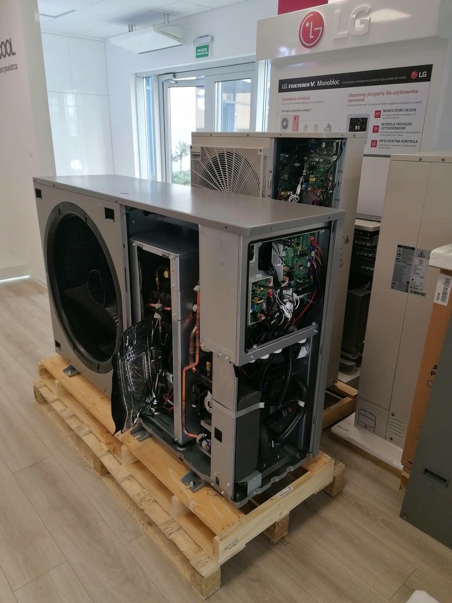 Rozpakowana pompa ciepła LG Therma V Monobloc na palecie w jasnym wnętrzu. Widoczne wewnętrzne komponenty i elektronika. Plakat reklamowy LG w tle.