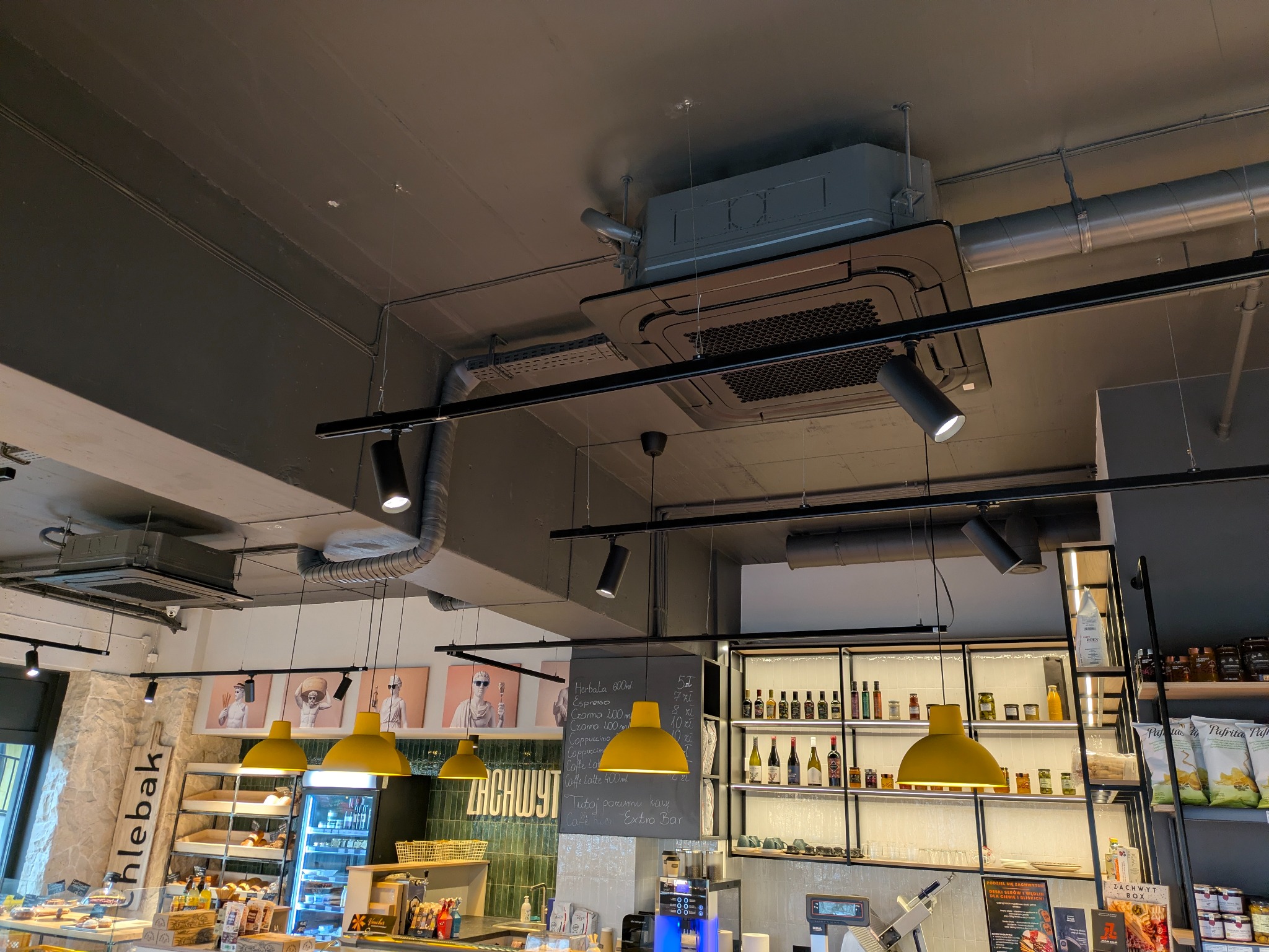 Sufit lokalu gastronomicznego z zamontowaną kasetą klimatyzacyjną, widoczne rury wentylacyjne, oświetlenie punktowe i wiszące lampy. Ujęcie z dołu.