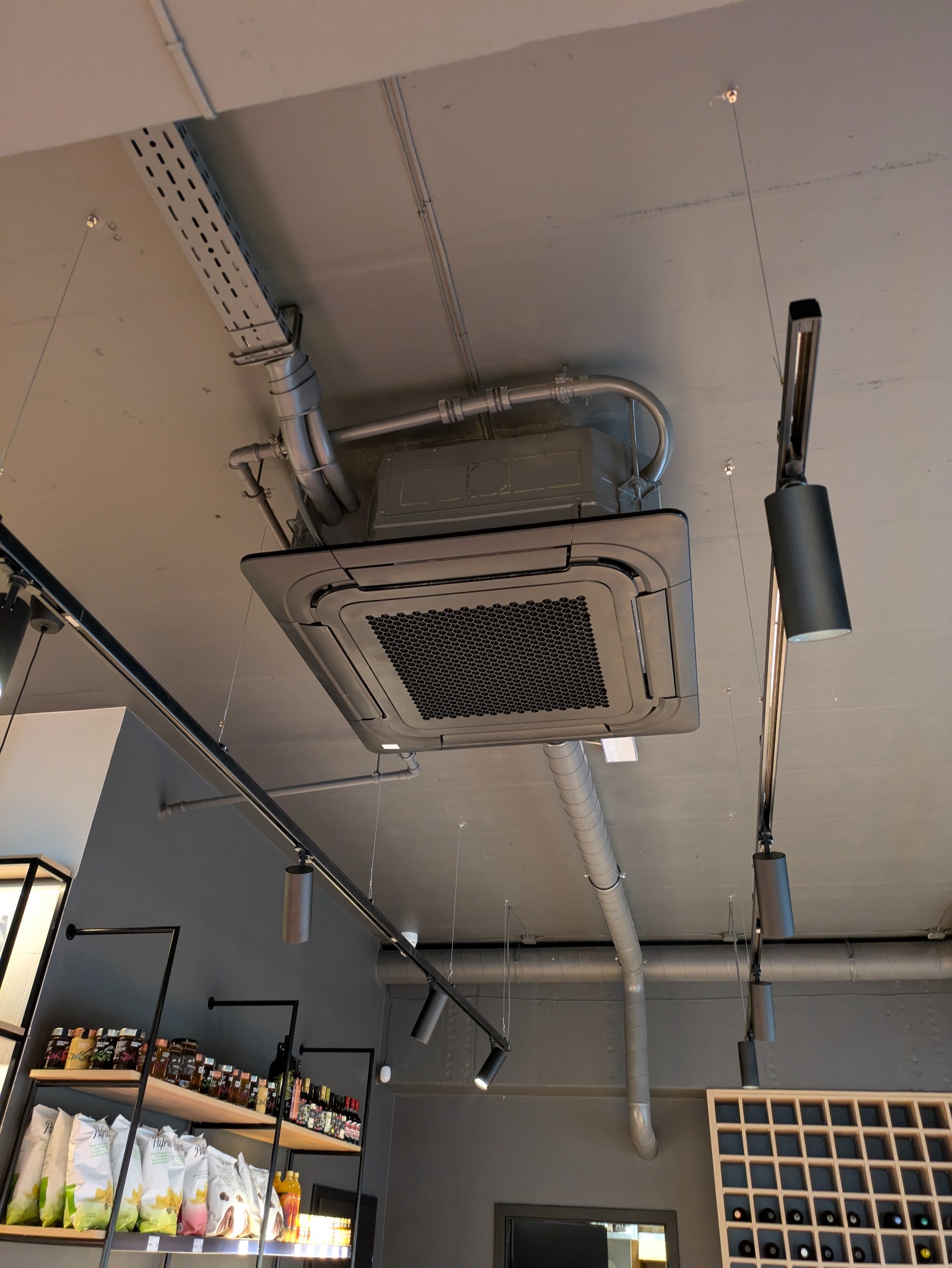 Sufitowa klimatyzacja kasetonowa w szarej tonacji, widoczne rury instalacyjne i oświetlenie punktowe. Nowoczesny system klimatyzacji w lokalu komercyjnym.