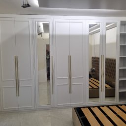 PPHU Prostyle Marcin Jankowski - Biała szafa wnękowa z lustrami i złotymi uchwytami, wbudowana w ścianę, obok widoczny fragment łóżka z beżowym zagłówkiem.