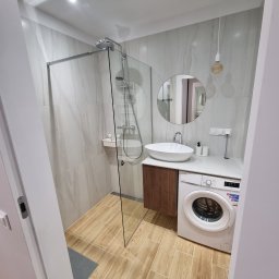 PPHU Prostyle Marcin Jankowski - Nowoczesna łazienka z kabiną prysznicową, okrągłym lustrem nad umywalką i pralką pod blatem. Beżowe płytki imitujące drewno na podłodze i marmur na ścianach.