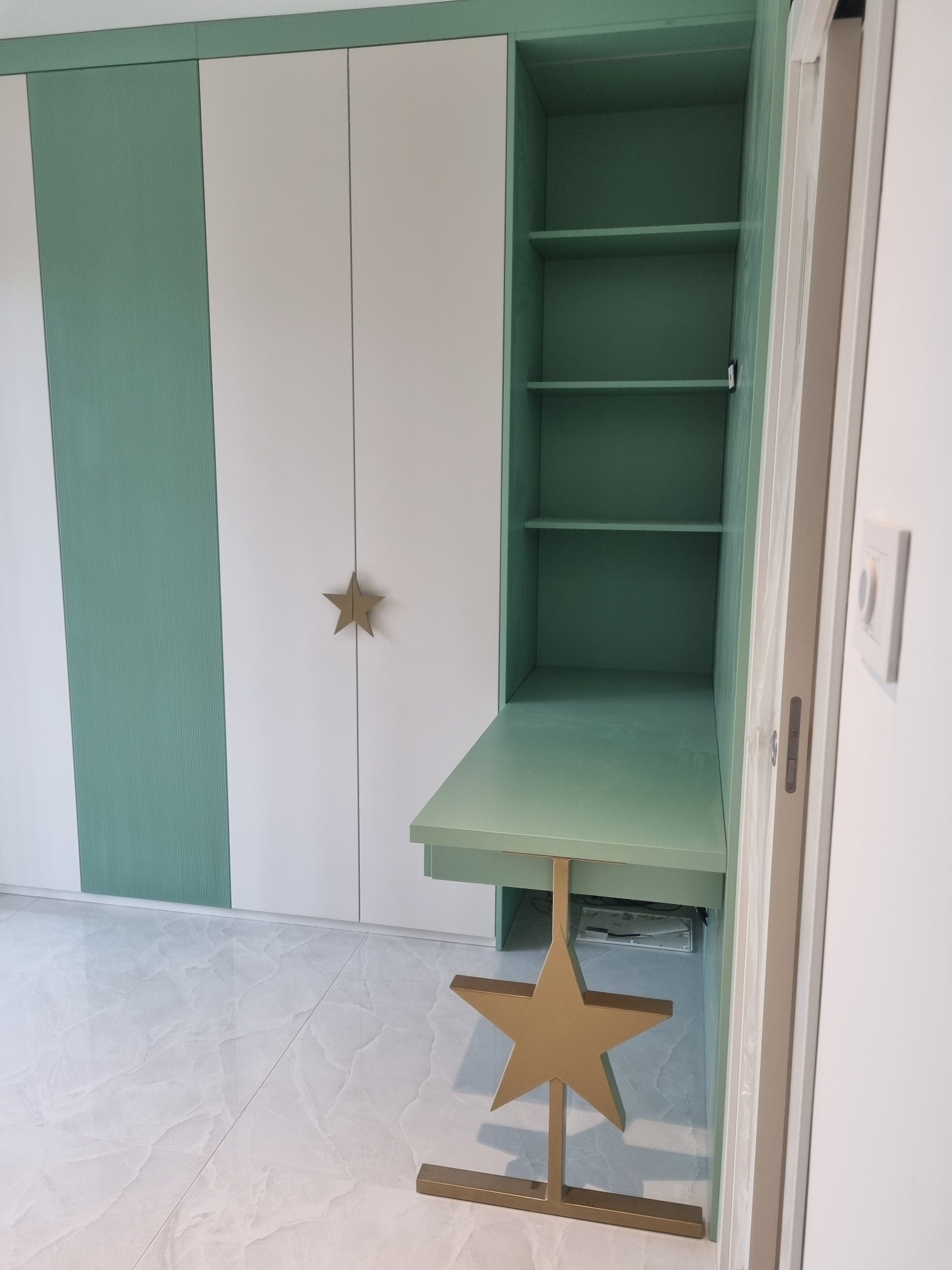 Biało-zielona szafa z biurkiem w pokoju dziecięcym. Drzwiczki szafy zdobią złote gwiazdki, a biurko wspiera konstrukcja w kształcie gwiazdy.