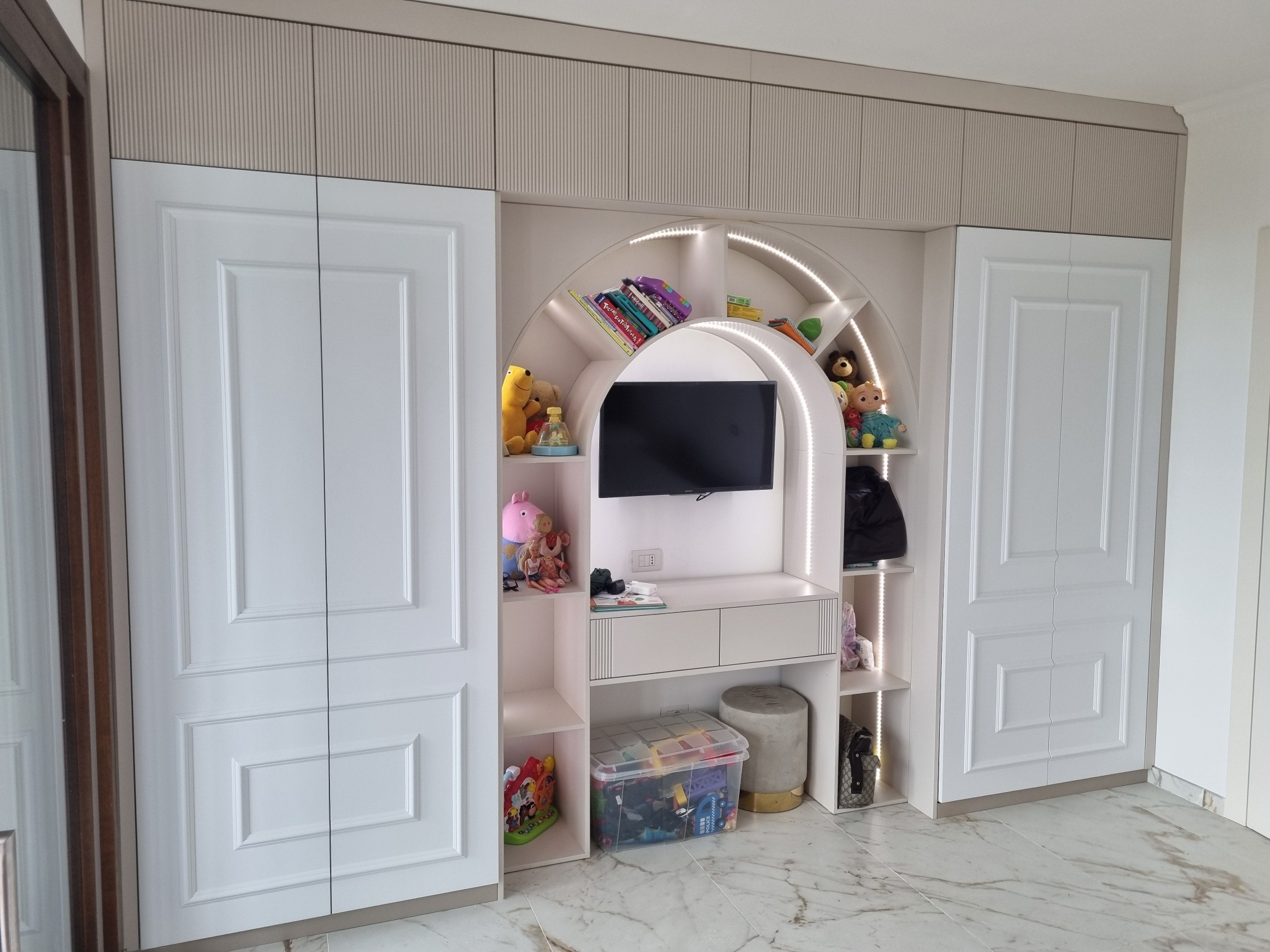Biała szafa wnękowa z półkami w kształcie łuku, podświetlona LED, z zabawkami i telewizorem, na marmurowej podłodze. Klasyczne frezowane fronty.