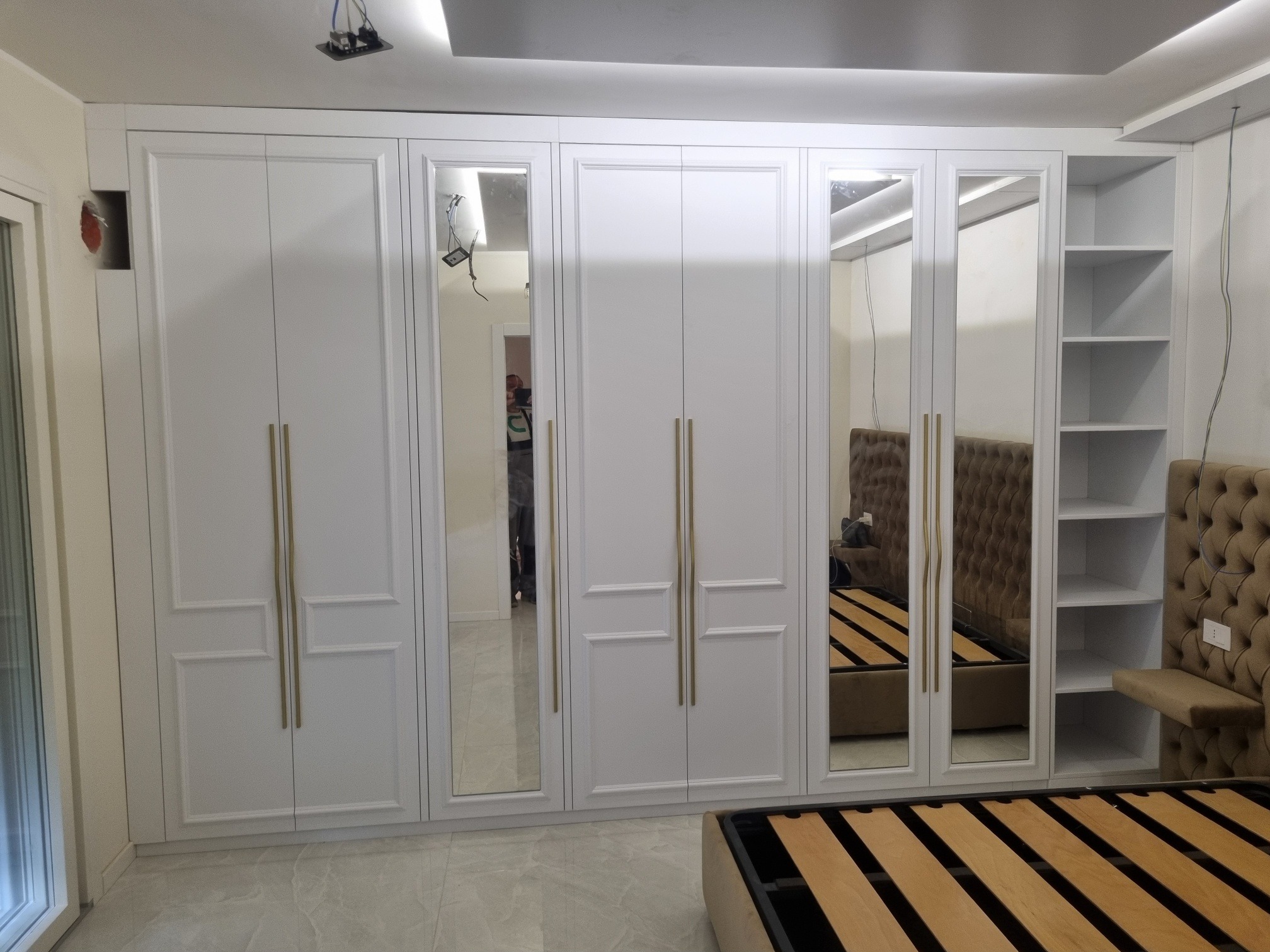 Biała szafa wnękowa z lustrami i złotymi uchwytami, wbudowana w ścianę, obok widoczny fragment łóżka z beżowym zagłówkiem.