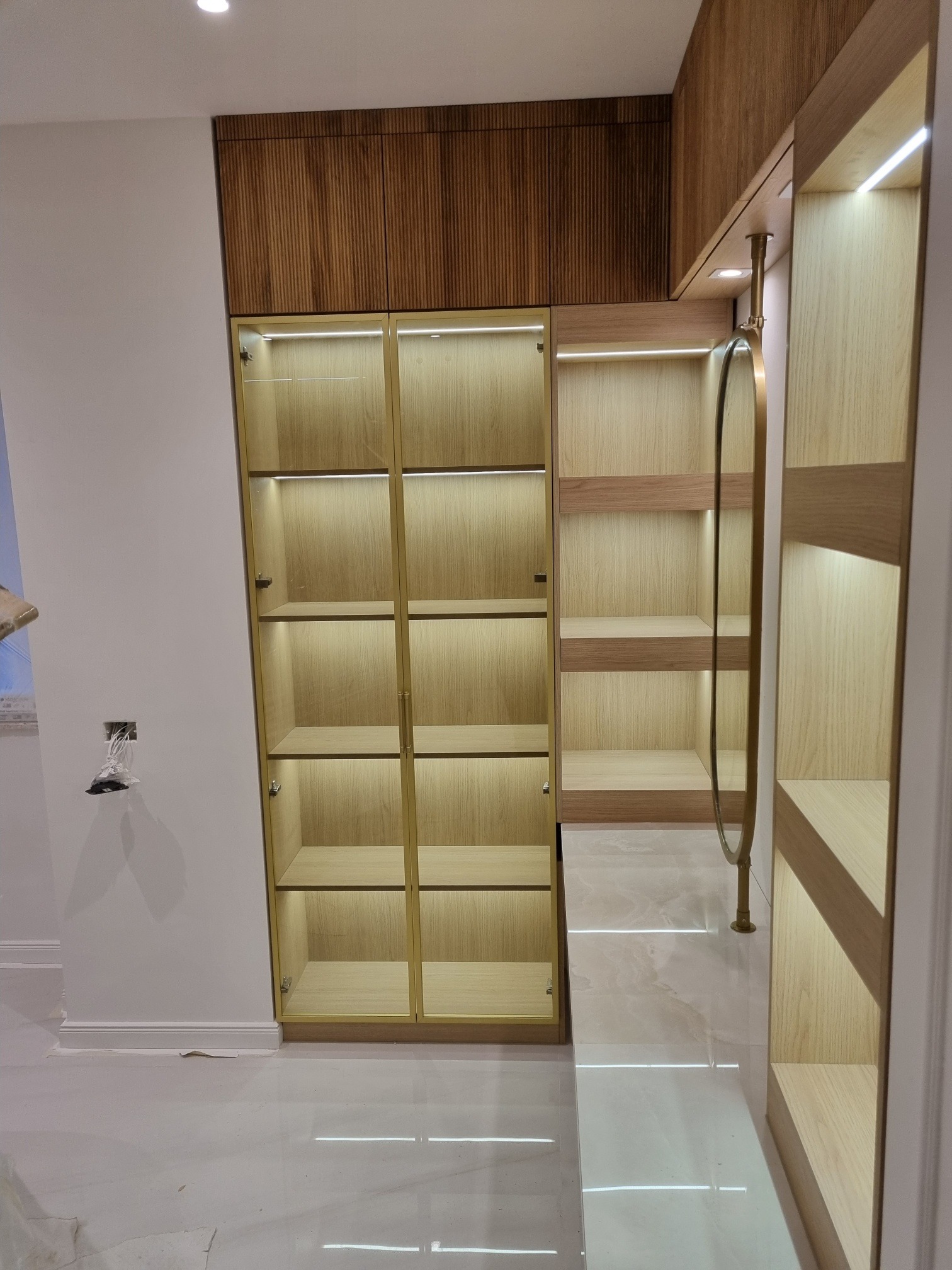 Wnętrze szafy z oświetleniem LED, drewnianymi półkami i złotymi ramami szklanych drzwi. Nad szafą widoczna drewniana zabudowa z żłobieniami.