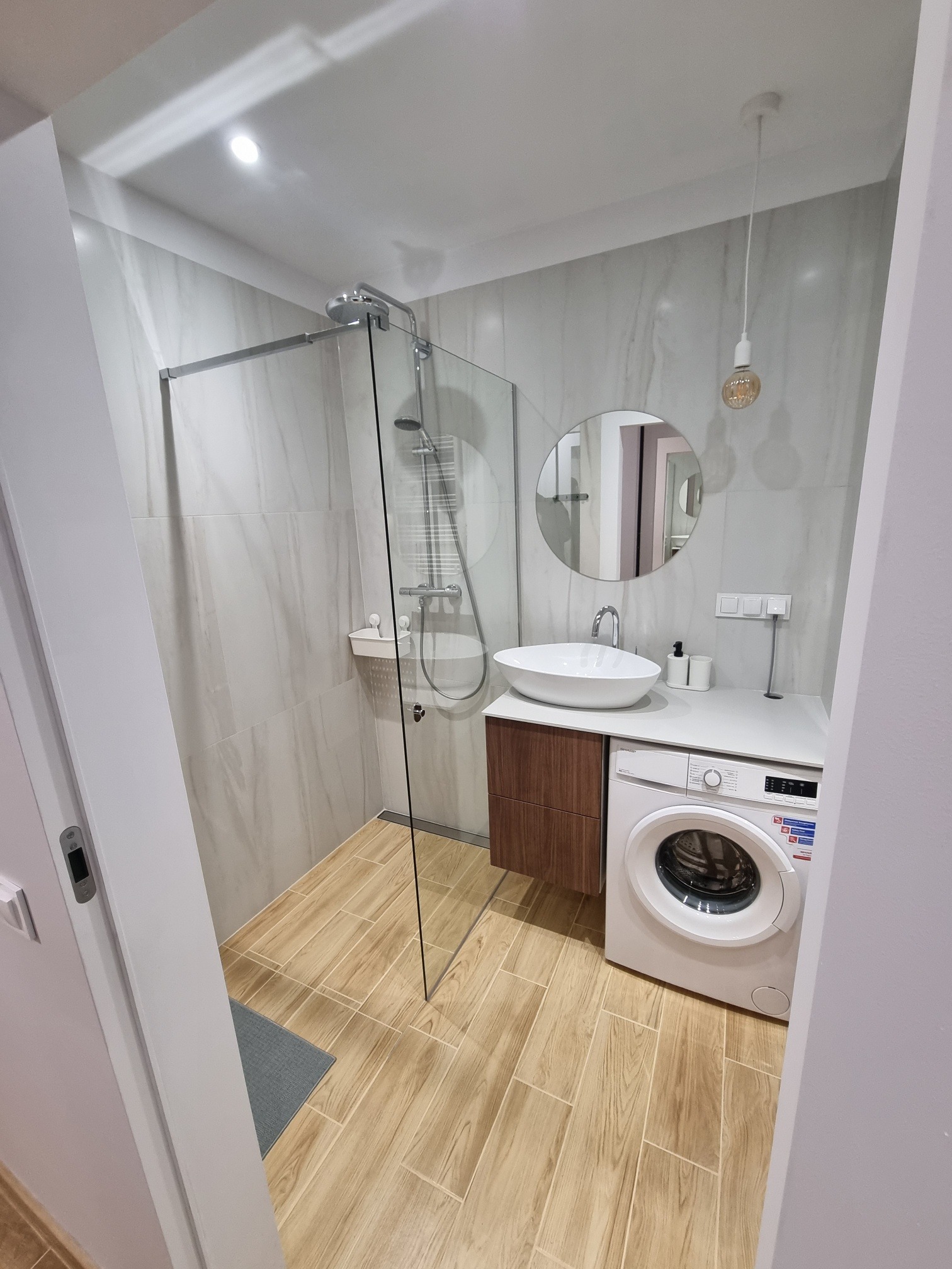 Nowoczesna łazienka z kabiną prysznicową, okrągłym lustrem nad umywalką i pralką pod blatem. Beżowe płytki imitujące drewno na podłodze i marmur na ścianach.