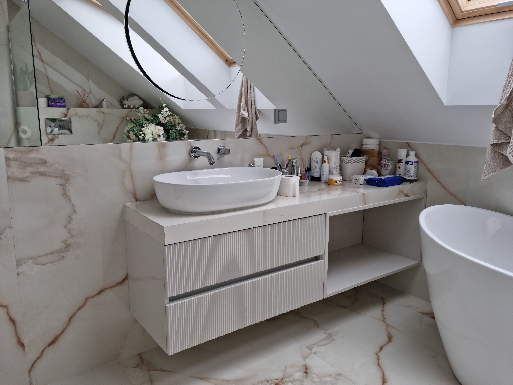 Nowoczesna łazienka na poddaszu z marmurową ścianą i podłogą. Biała umywalka nablatowa i szafka z frezowanymi frontami. Okrągłe lustro odbija okno dachowe.