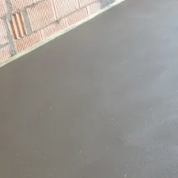 Świeżo wylana, gładka, szara posadzka jastrychowa przylegająca do ceglanej ściany z widoczną izolacją. Widoczne włókna zbrojeniowe w masie.