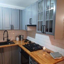 WYKOŃCZENI.PL Paweł Magusiewicz - Szara kuchnia z drewnianym blatem, białymi kaflami i czarną armaturą, oświetlona świecą, z widoczną płytą grzewczą i elementami dekoracyjnymi.