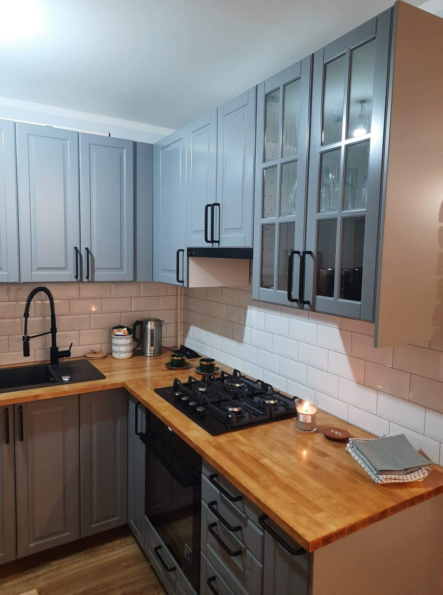 Szara kuchnia z drewnianym blatem, białymi kaflami i czarną armaturą, oświetlona świecą, z widoczną płytą grzewczą i elementami dekoracyjnymi.