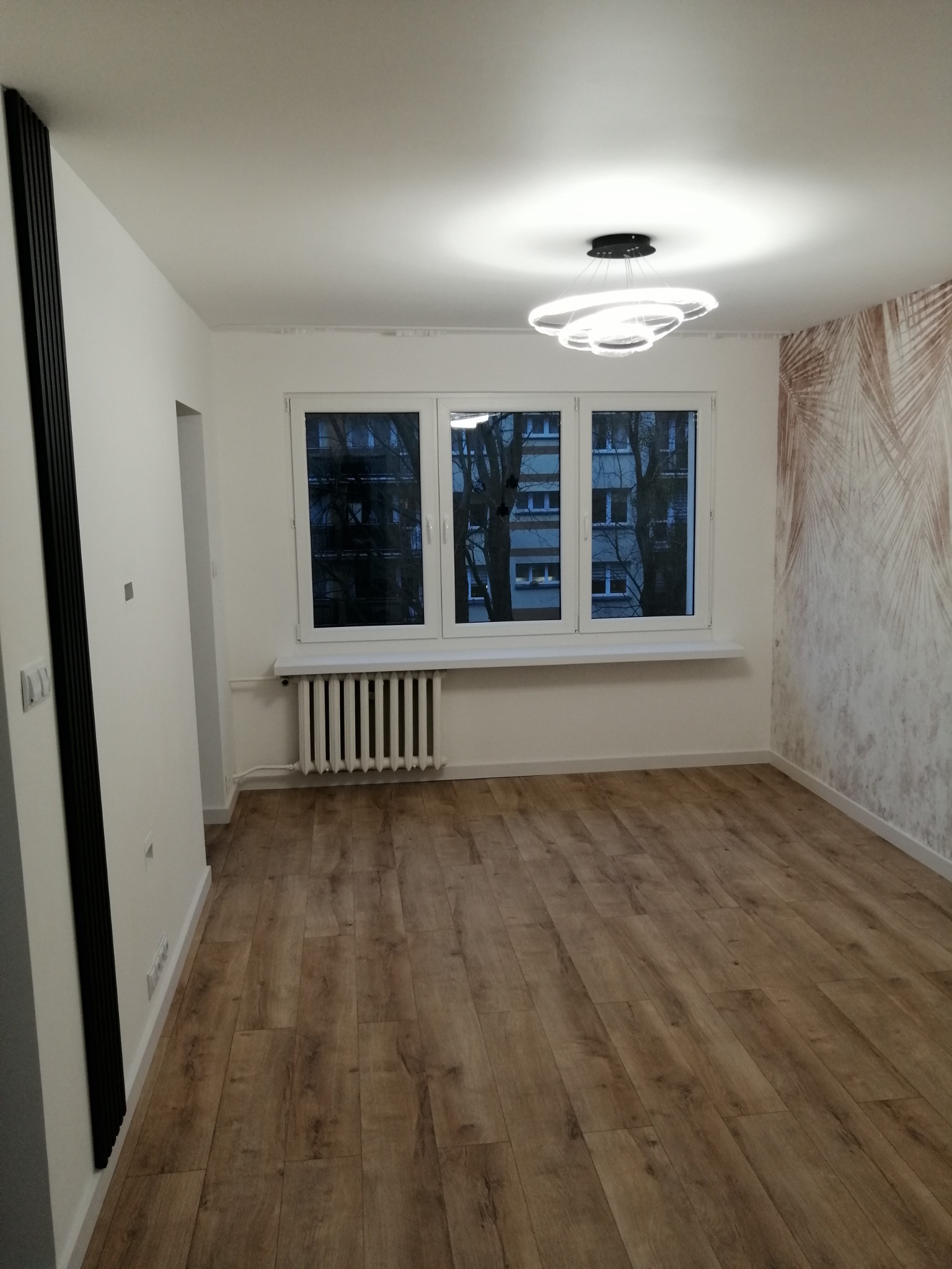 Pusty pokój po odświeżeniu: drewnopodobne panele podłogowe, białe ściany z akcentem tapety w liście palmowe, nowoczesna lampa sufitowa i grzejnik pod trzema oknami.