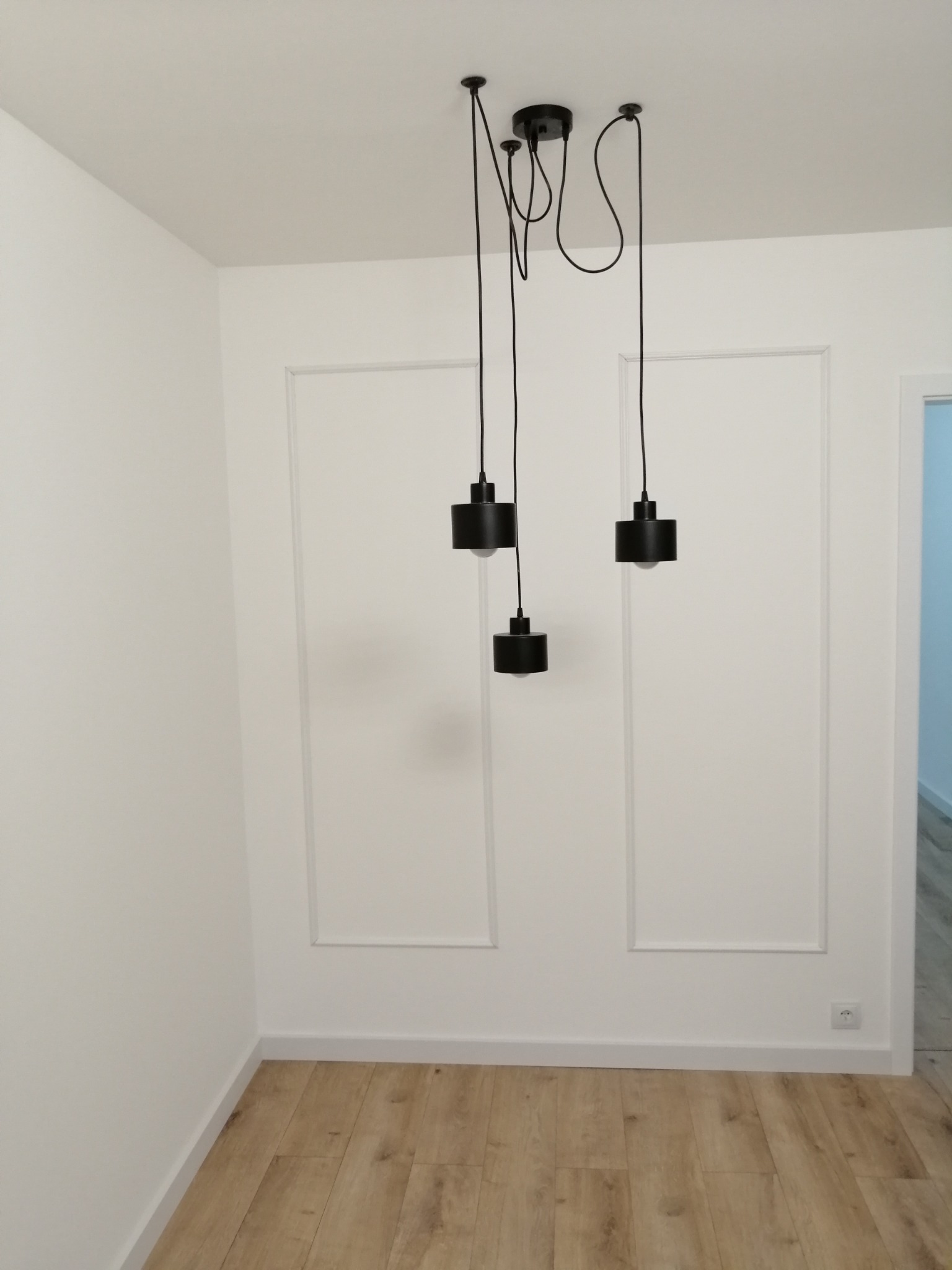 Nowoczesne, czarne lampy wiszące z odsłoniętymi żarówkami, zwisające z sufitu w białym pokoju z panelami podłogowymi i dekoracyjnymi listwami na ścianach.