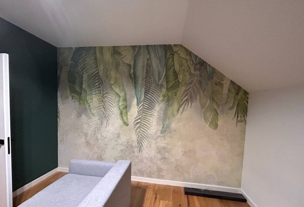 Pokój na poddaszu z fototapetą w motywy tropikalnych liści, ciemnozielona ściana kontrastuje z jasną tapetą i drewnianą podłogą, widoczny fragment szarej sofy.