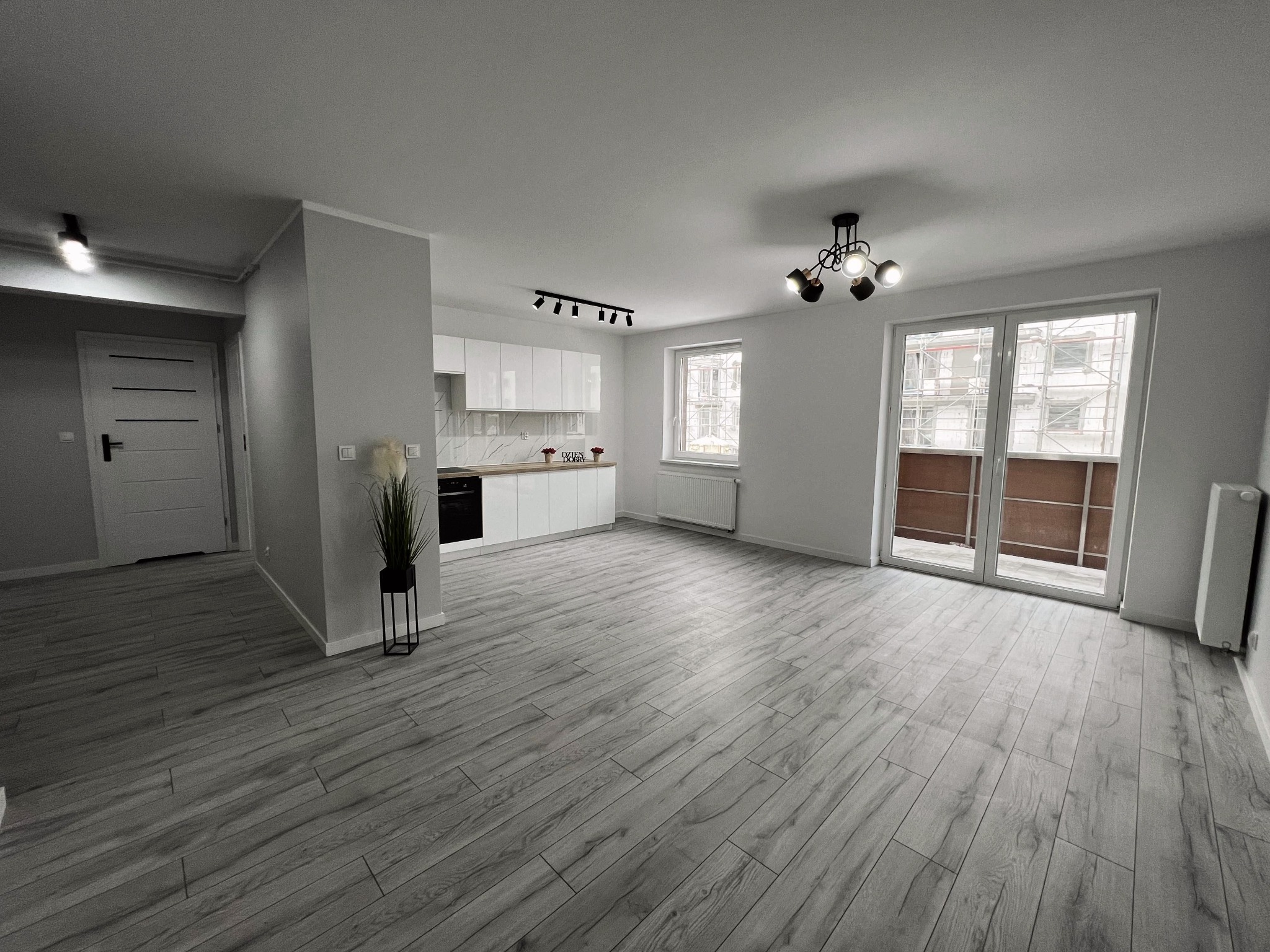 Jasne, przestronne wnętrze po remoncie: szare panele podłogowe imitujące drewno, biała zabudowa kuchenna z drewnianym blatem, nowoczesne oświetlenie sufitowe i widok na rusztowania za oknem...