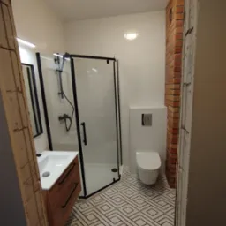 Nowoczesna łazienka z kabiną prysznicową z czarnymi ramami, umywalką z drewnianą szafką, toaletą podwieszaną i geometryczną podłogą, fragment ściany z cegły oraz surowy otwór drzwiowy.