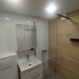 AWbud - Nowoczesna łazienka z kabiną prysznicową typu walk-in, białą umywalką z szafką i lustrem. Ściany wyłożone białymi i drewnopodobnymi płytkami. Minimalistyczny design.