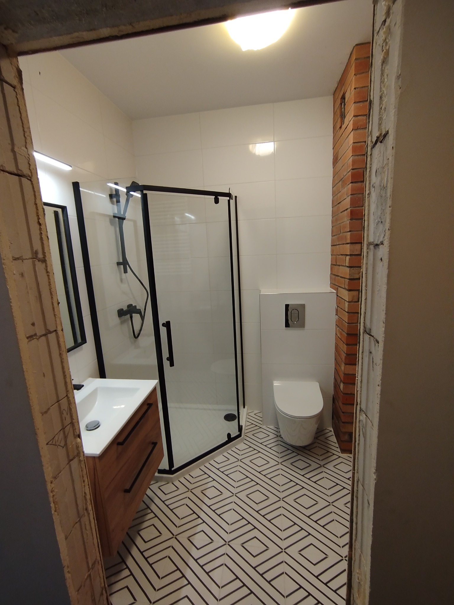Nowoczesna łazienka z kabiną prysznicową z czarnymi ramami, umywalką z drewnianą szafką, toaletą podwieszaną i geometryczną podłogą, fragment ściany z cegły oraz surowy otwór drzwiowy.