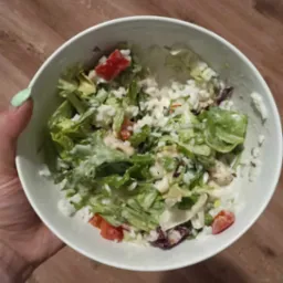 Sałatka z ryżem, pomidorami i sosem w białej misce trzymanej w dłoni z jasnozielonym paznokciem na tle drewnopodobnej powierzchni.