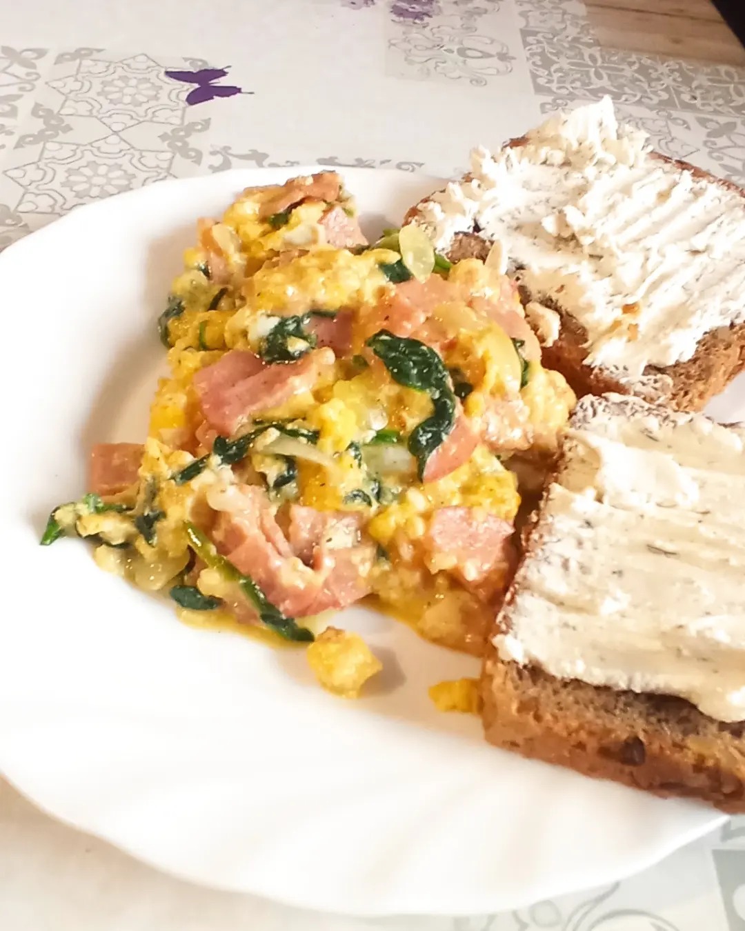 Śniadanie na białym talerzu: jajecznica ze szpinakiem i szynką obok dwóch kromek chleba razowego posmarowanych kremowym serkiem.