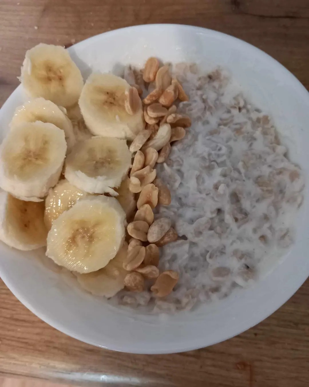 Owsianka z bananem w plasterkach i orzeszkami ziemnymi na białym talerzu, na drewnianym stole.