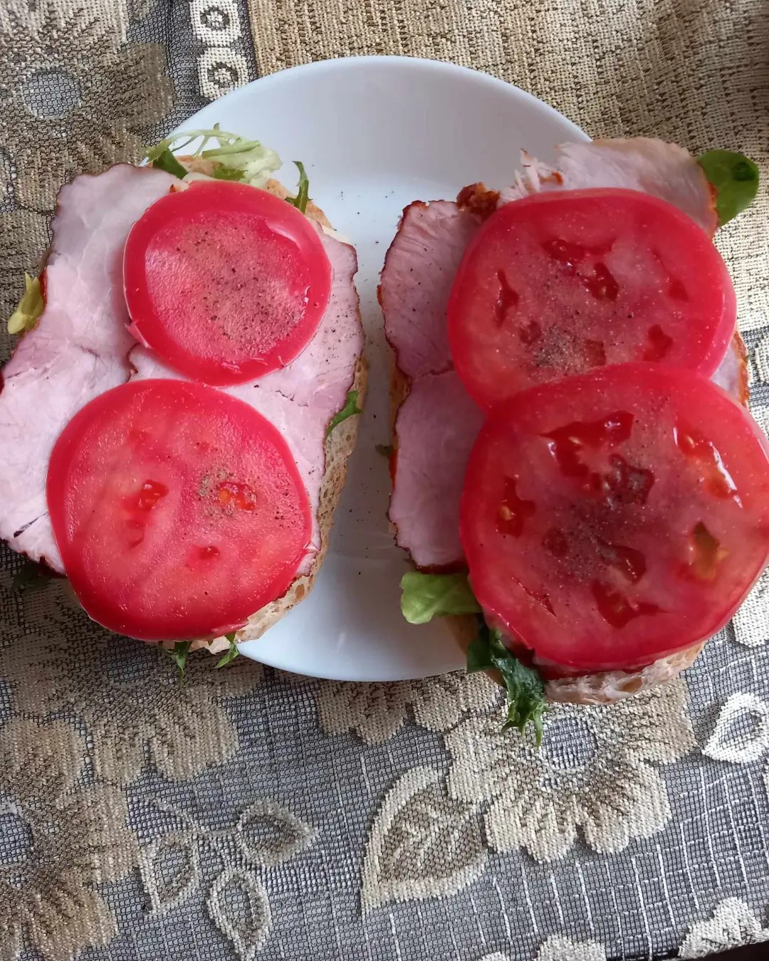 Dwa kanapki na białym talerzu z szynką, sałatą i dwoma plastrami pomidora na każdym, posypane pieprzem, na wzorzystym obrusie.