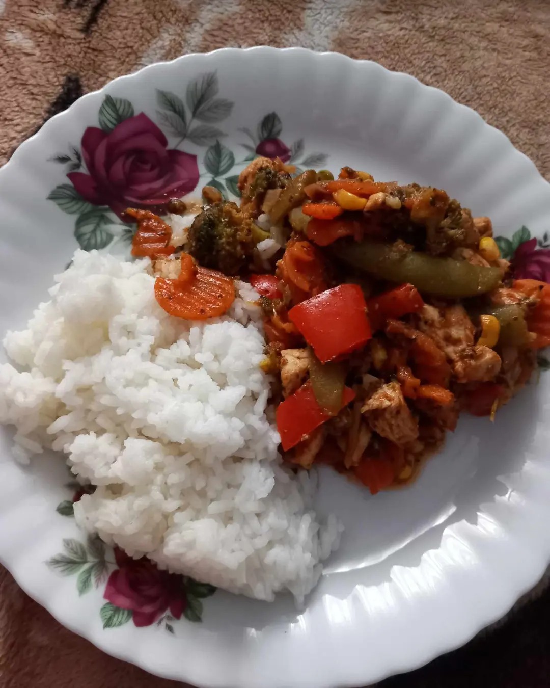 Porcja ryżu z warzywami i kurczakiem na białym talerzu z motywem róż, ułożona na beżowym tle.