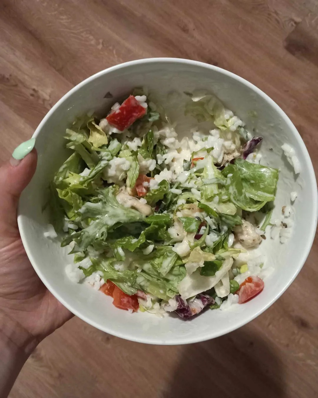 Sałatka z ryżem, pomidorami i sosem w białej misce trzymanej w dłoni z jasnozielonym paznokciem na tle drewnopodobnej powierzchni.