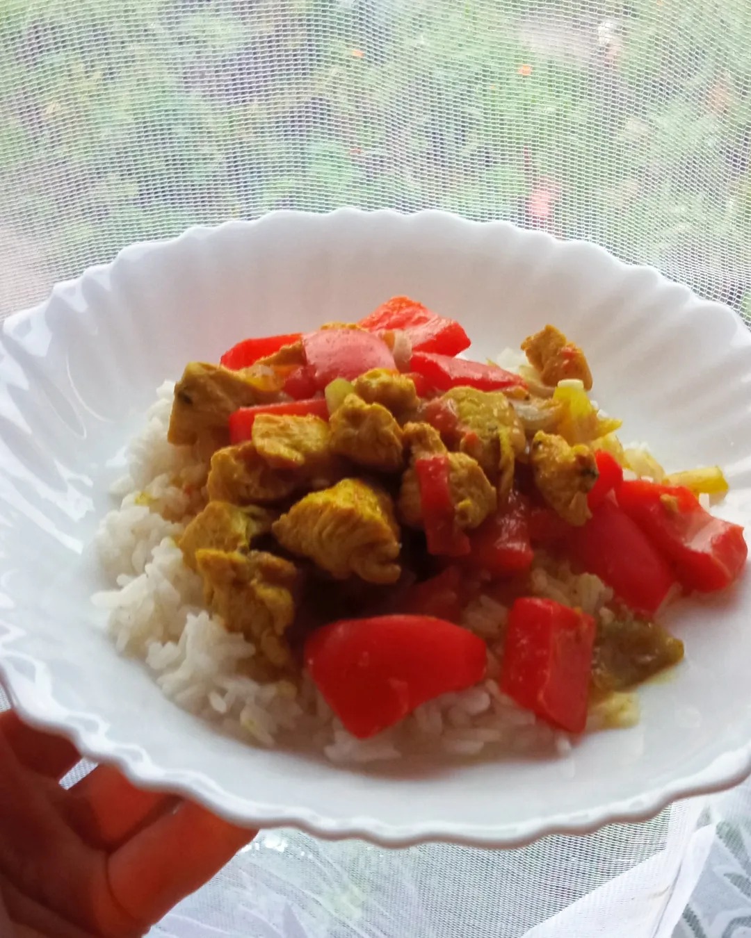 Porcja ryżu z kurczakiem curry i czerwoną papryką na białym talerzu trzymanym w dłoni, na tle firanki okiennej.