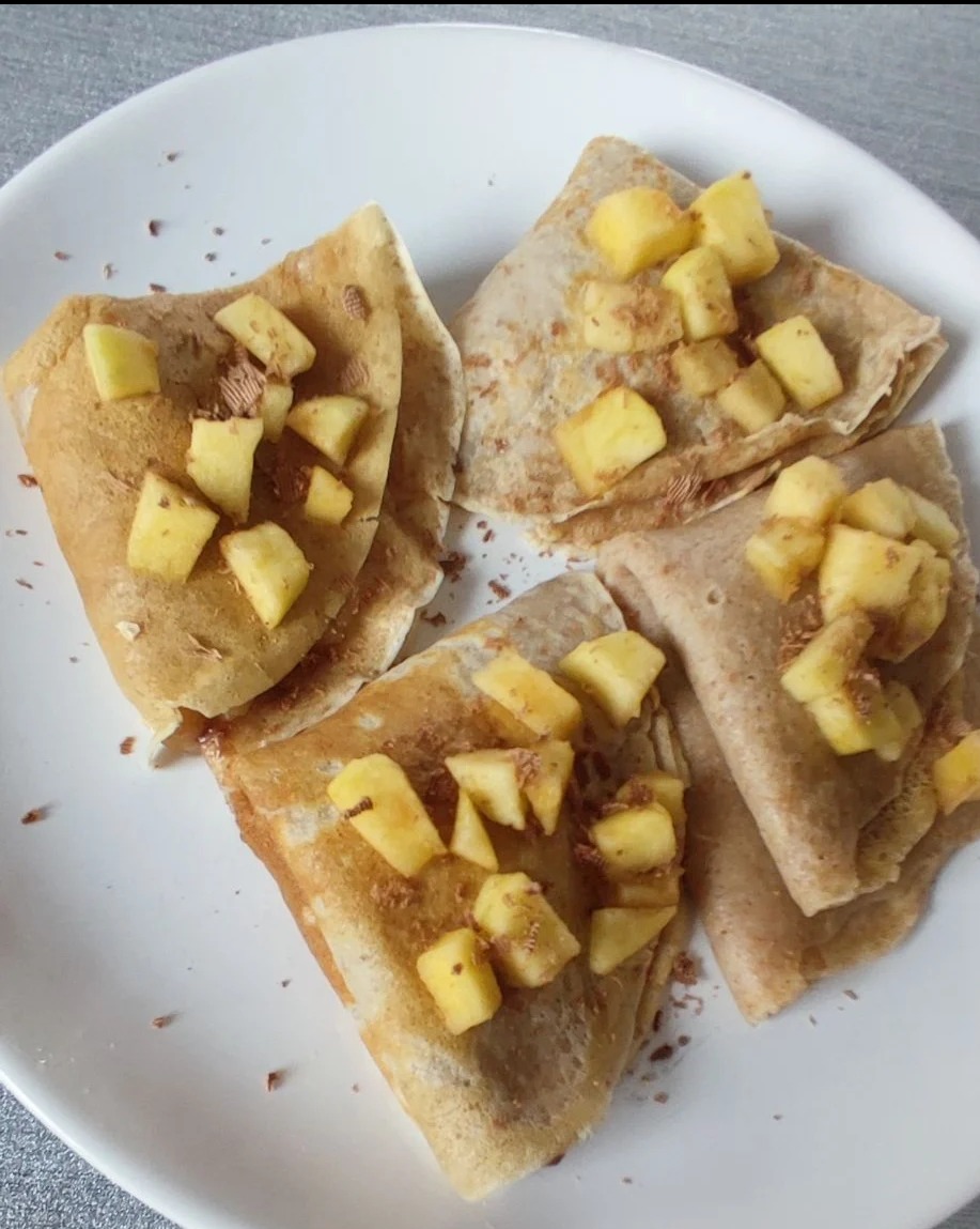 Trzy naleśniki gryczane złożone w trójkąty, wypełnione kostkami jabłek i posypane startą czekoladą, ułożone na białym talerzu.