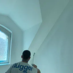 Mężczyzna w koszulce marki Fl&uuml;gger maluje ścianę natryskowo na biało, okno zabezpieczone folią.
