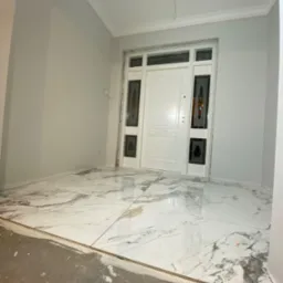 Nowoczesne wejście do domu z białymi ścianami, białymi drzwiami z ozdobnymi szybami i podłogą wyłożoną płytkami imitującymi biały marmur, widoczne ślady prac wykończeniowych na podłodze.