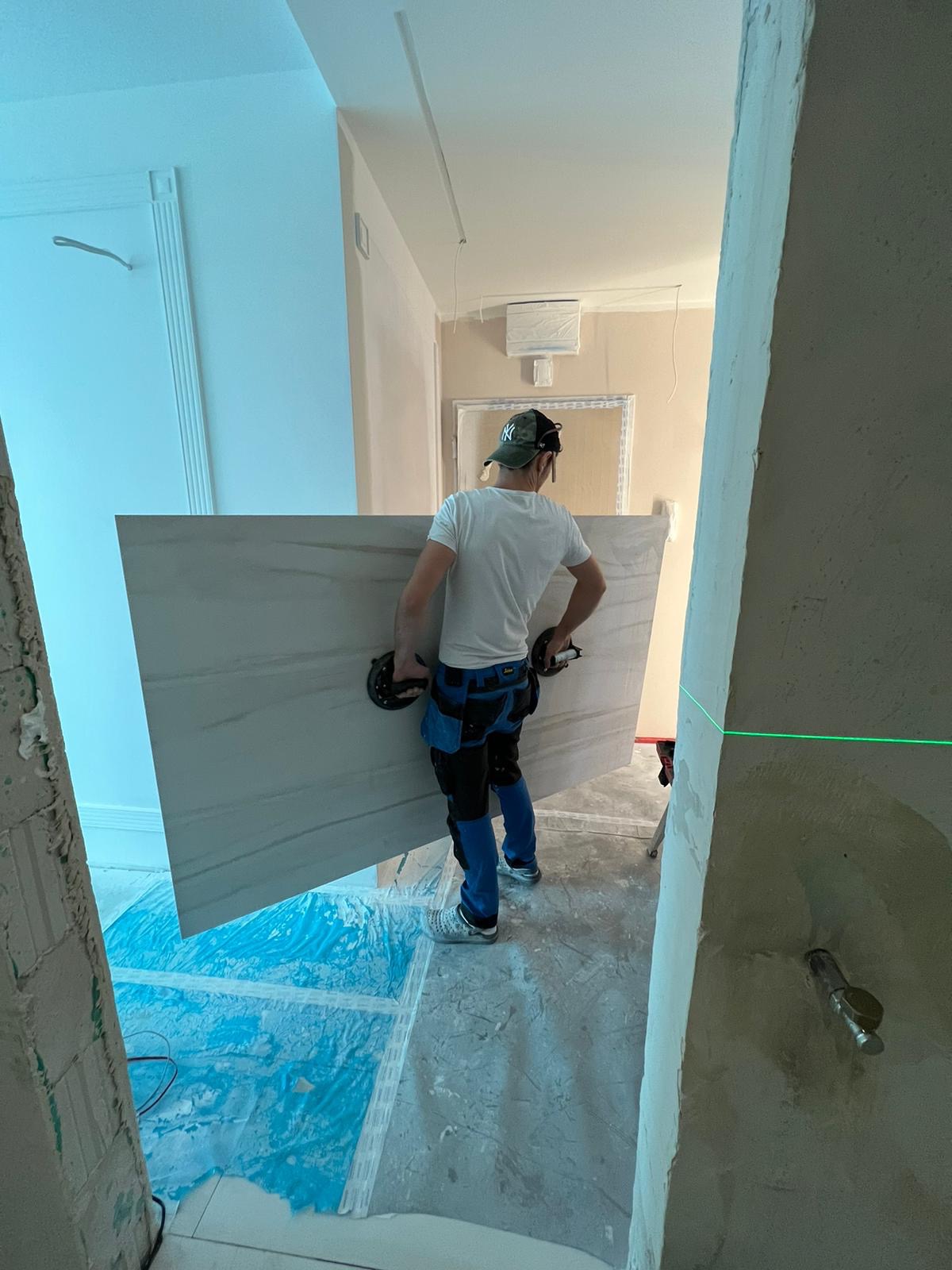 Pracownik w czapce niesie dużą płytę imitującą marmur przy użyciu przyssawek, widoczny laserowy poziom na ścianie, podłoga zabezpieczona folią.