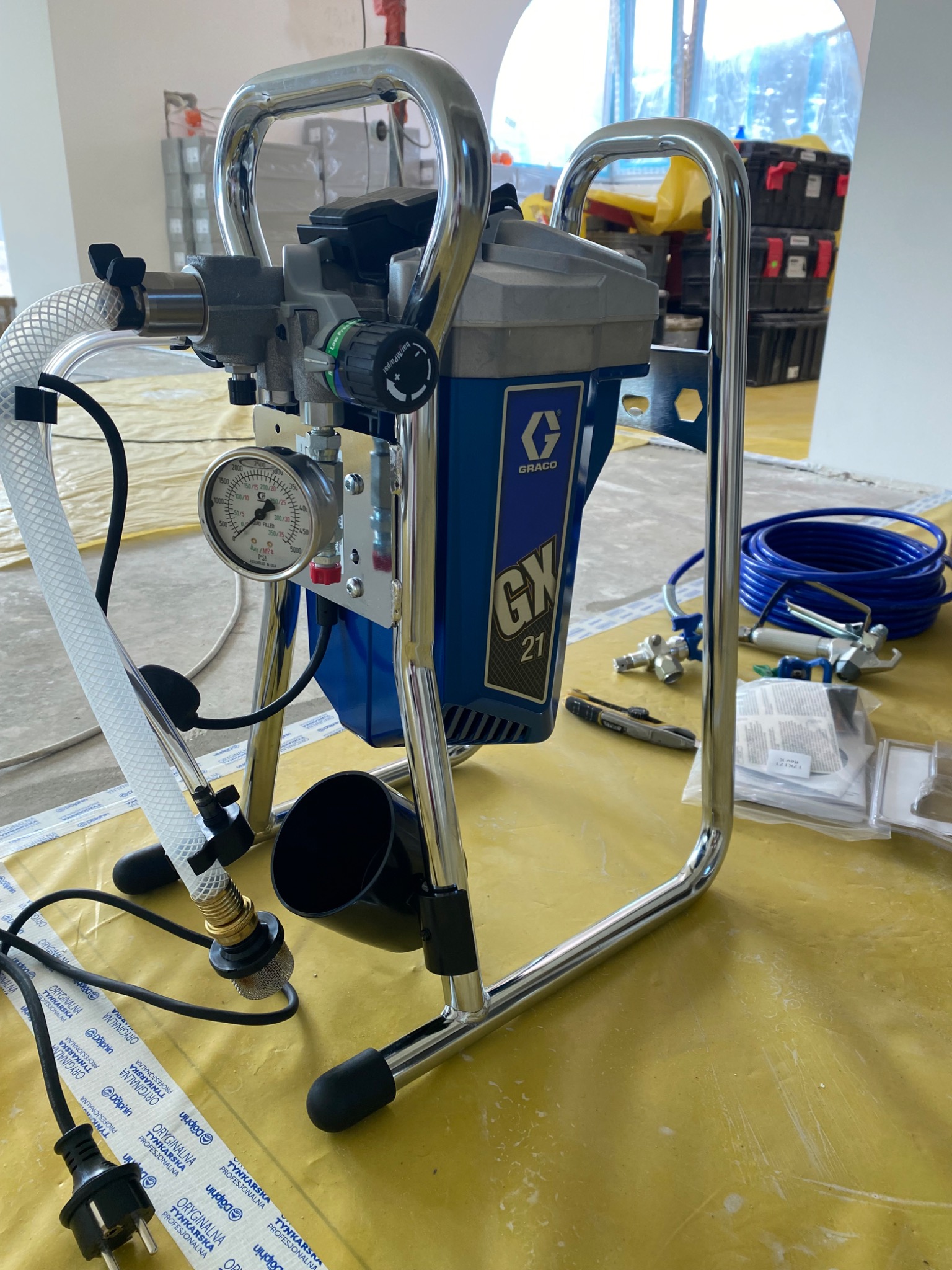 Profesjonalny agregat malarski Graco GX 21 na żółtej folii ochronnej, z widocznym manometrem i akcesoriami, w tle ścianka działowa i skrzynki narzędziowe.