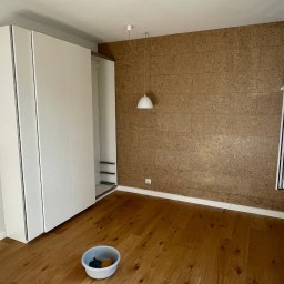 Oskar Kubiak firma ogólnobudowlana - Pokój po remoncie: drewniana podłoga, ściana wyłożona korkiem, biała szafa w zabudowie, wisząca lampa i otwarte okno z widokiem na zewnątrz; na podłodze wiadro z narzędziami.