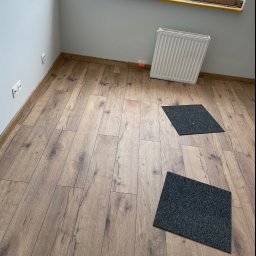 Oskar Kubiak firma ogólnobudowlana - Wykończone wnętrze pokoju z panelami podłogowymi o wzorze drewna, białymi ścianami, grzejnikiem pod oknem i dwoma szarymi dywanikami.