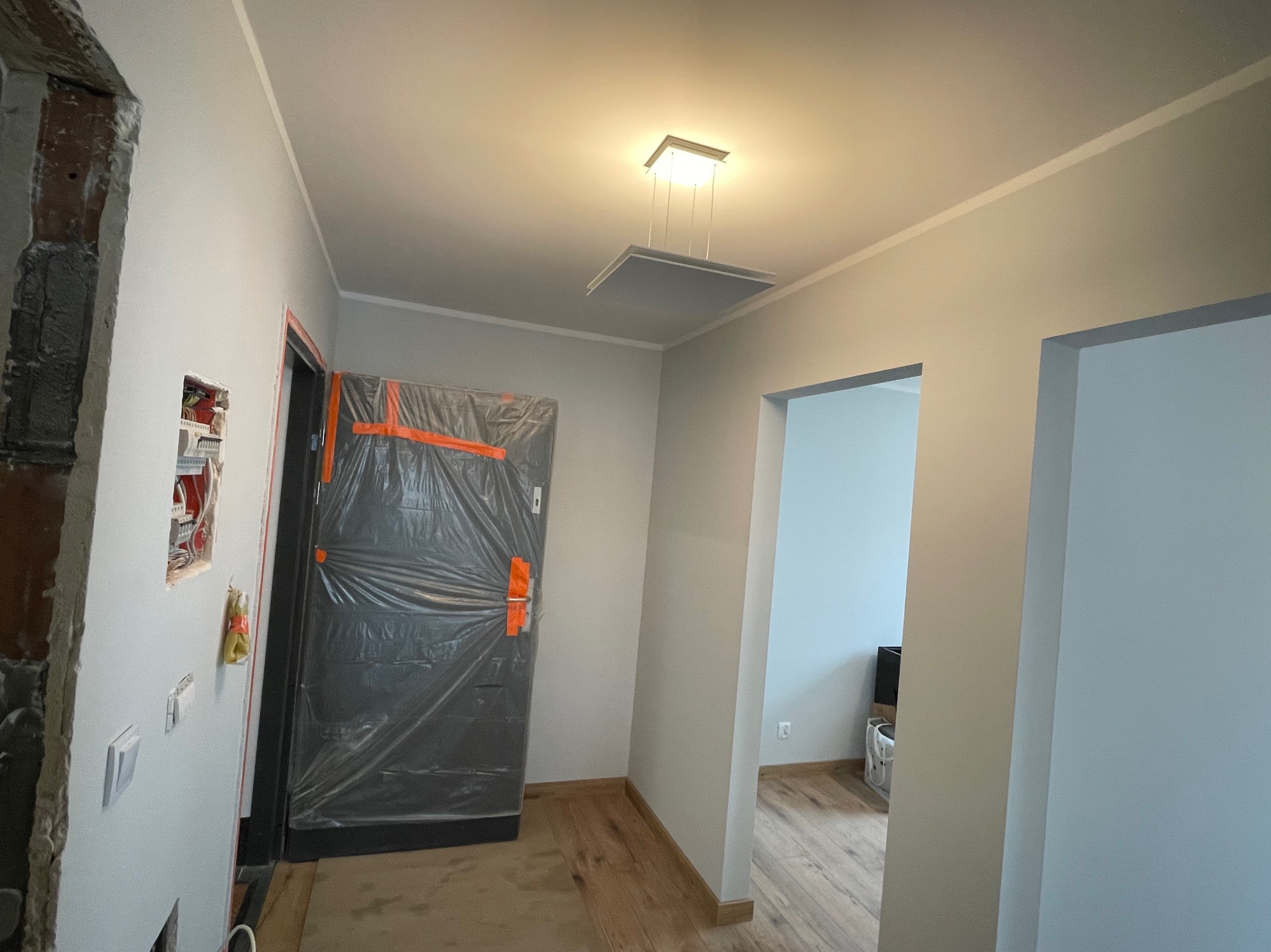 Korytarz w trakcie prac wykończeniowych: ściany pomalowane na szaro, widoczna instalacja elektryczna, drzwi zabezpieczone folią ochronną, sufit z nowoczesnym oświetleniem.