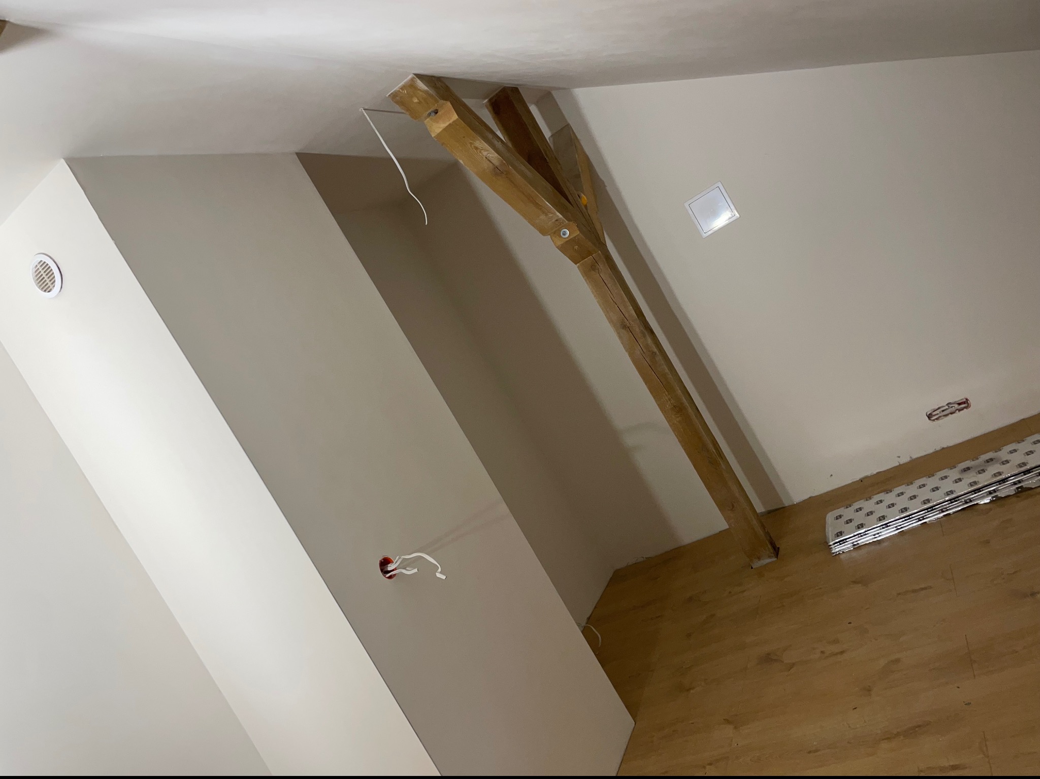 Skośne poddasze po pracach wykończeniowych; widoczne pomalowane ściany w odcieniach beżu, drewniane belki konstrukcyjne, wentylacja, gniazdko elektryczne z wystającymi kablami oraz ułożona podłoga...