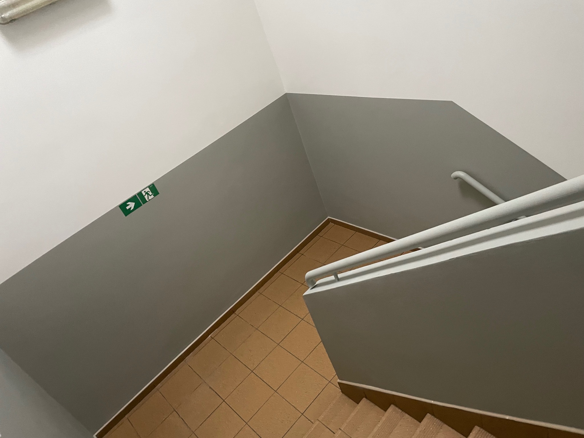 Klatka schodowa z pomalowanymi na szaro ścianami, brązowymi płytkami na podłodze i białymi poręczami, widok z góry, z zielonym znakiem ewakuacyjnym na ścianie.