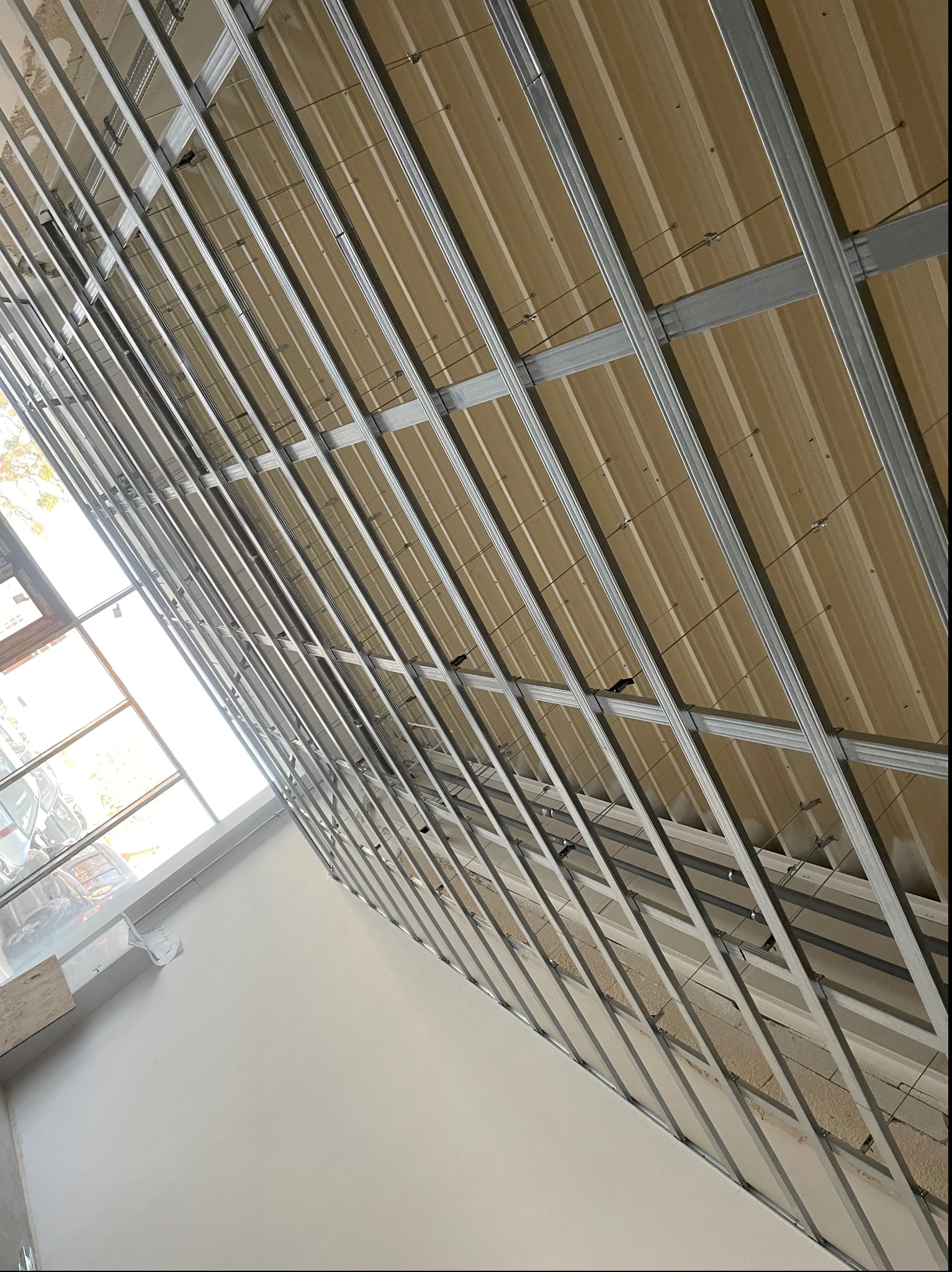 Montaż metalowej konstrukcji sufitu podwieszanego z widoczną izolacją, widok z dołu pod kątem, z oknem w tle.